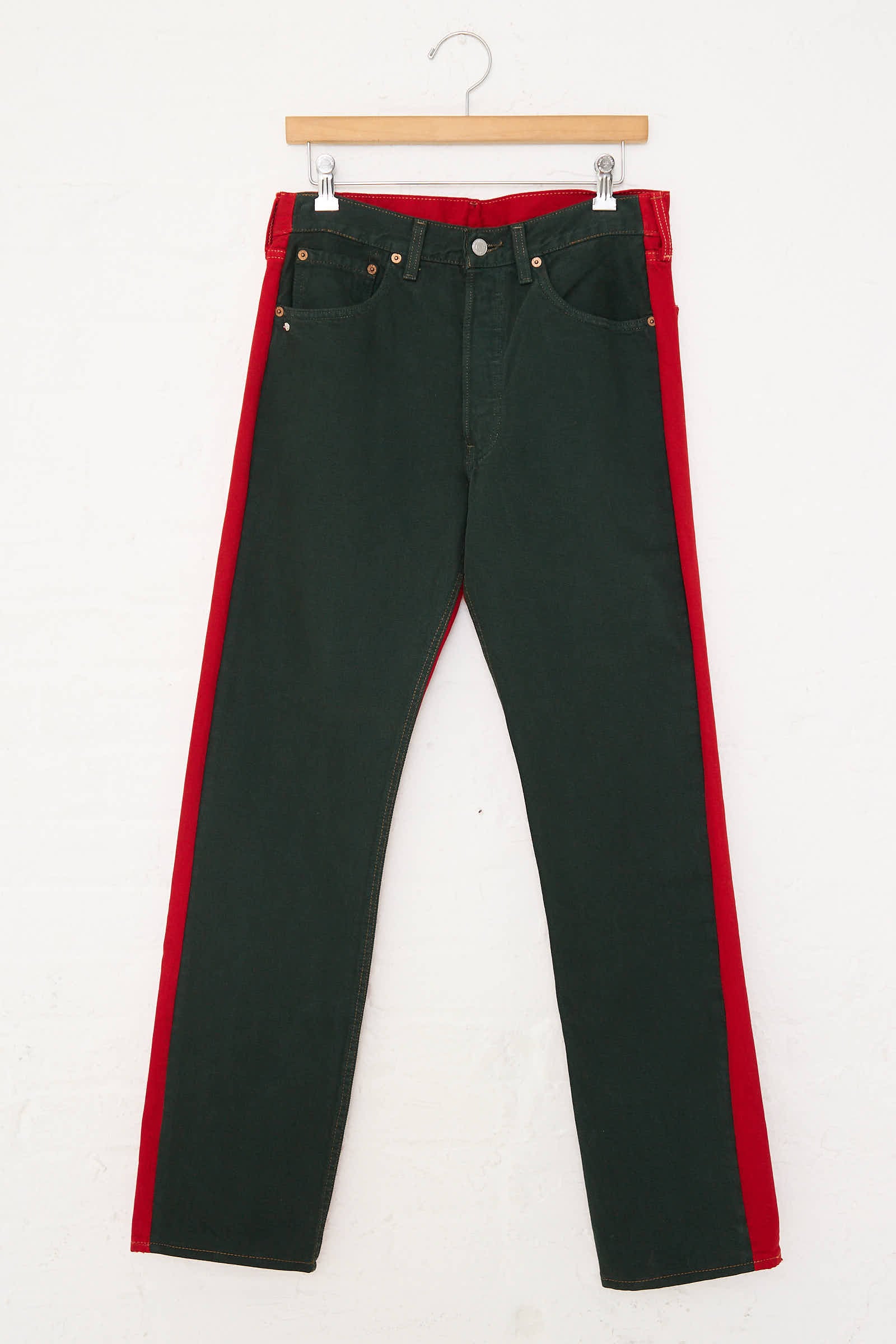 A pair of No. 73 Jeanspleatfront in Black/Red by Bless, featuring mid-waist design and red side panels, crafted from up-cycled vintage denim, hangs on a wooden hanger against a white background.