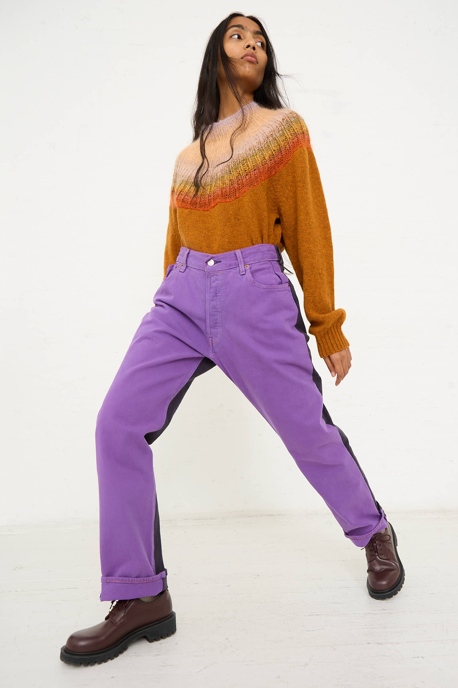 A person stands against a plain background, wearing a patterned sweater, Bless No. 73 Jeanspleatfront in Purple/Black with a relaxed fit, and brown boots. They are posing with one leg stretched forward.