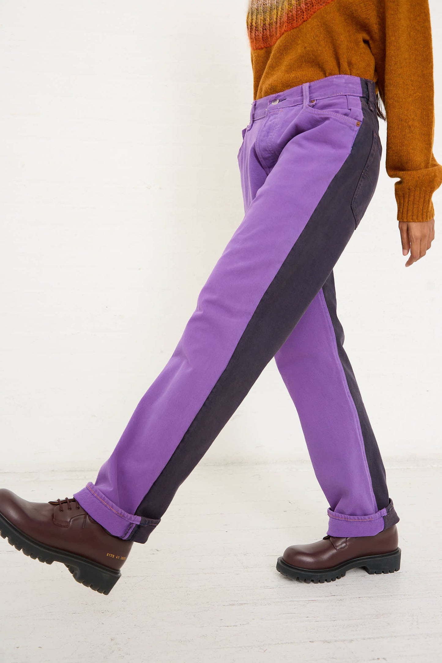 Side view of a person walking in No. 73 Jeanspleatfront in Purple/Black by Bless, complemented with brown boots, an orange sweater, and accentuated with up-cycled vintage denim for a unique touch.