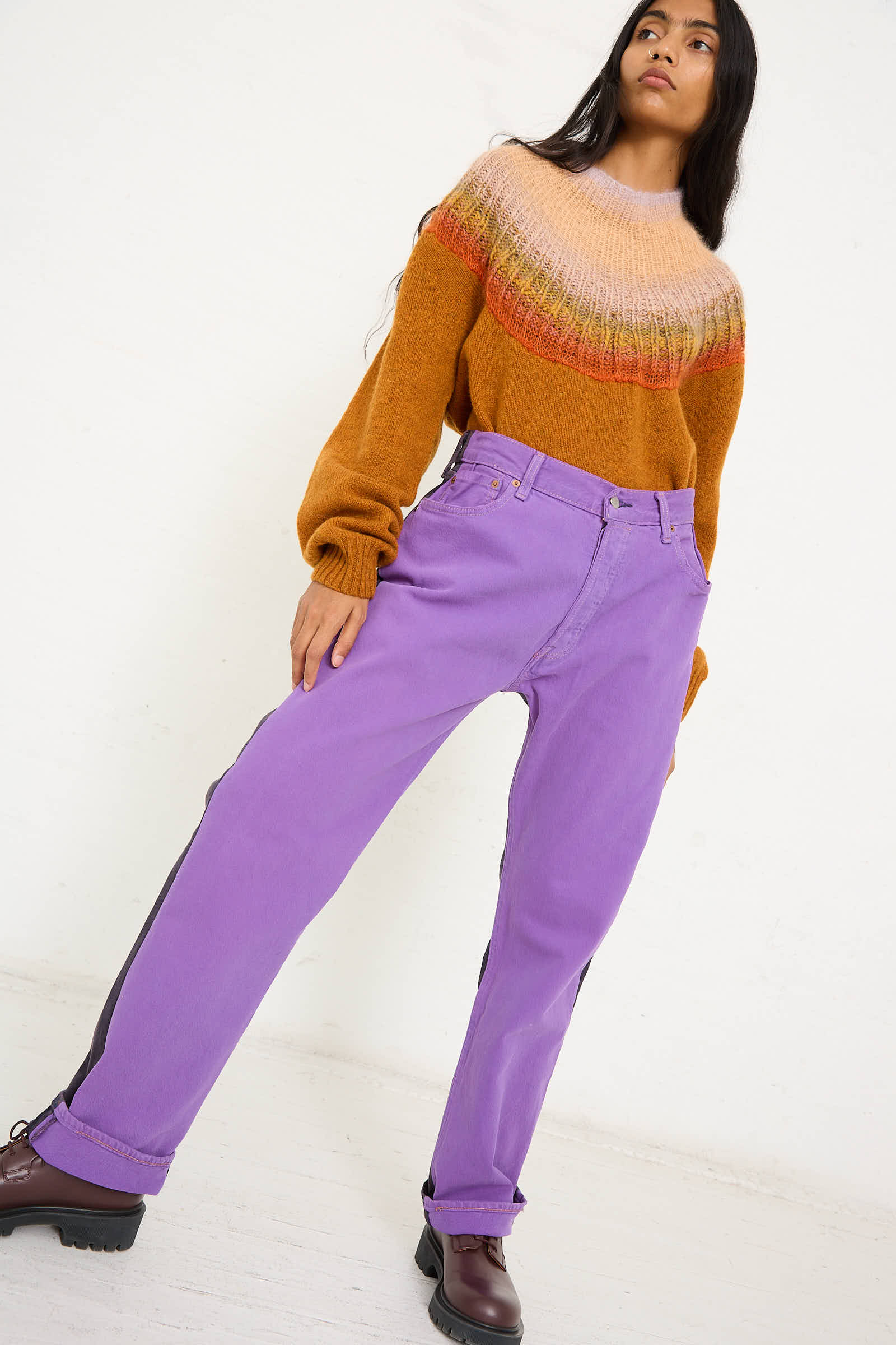 A person stands against a white background, wearing a multicolored sweater with hues of orange and pink, mid-waist relaxed-fit pants from Bless's No. 73 Jeanspleatfront collection in Purple/Black, and dark shoes.