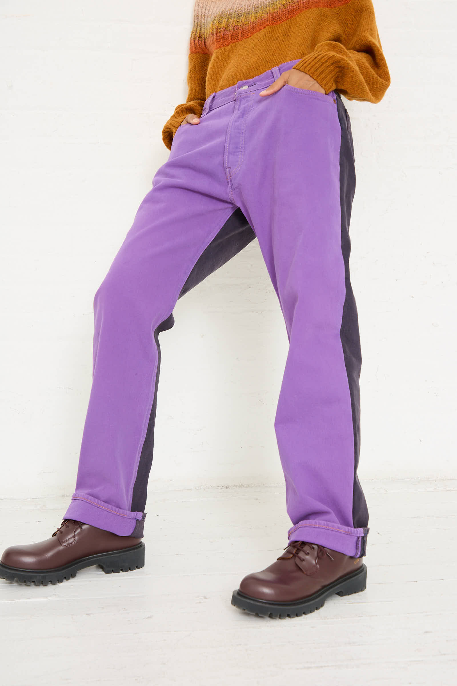 Person wearing Bless No. 73 Jeanspleatfront in Purple/Black, brown ankle boots, and a mustard orange sweater stands against a white background, showcasing relaxed fit style.