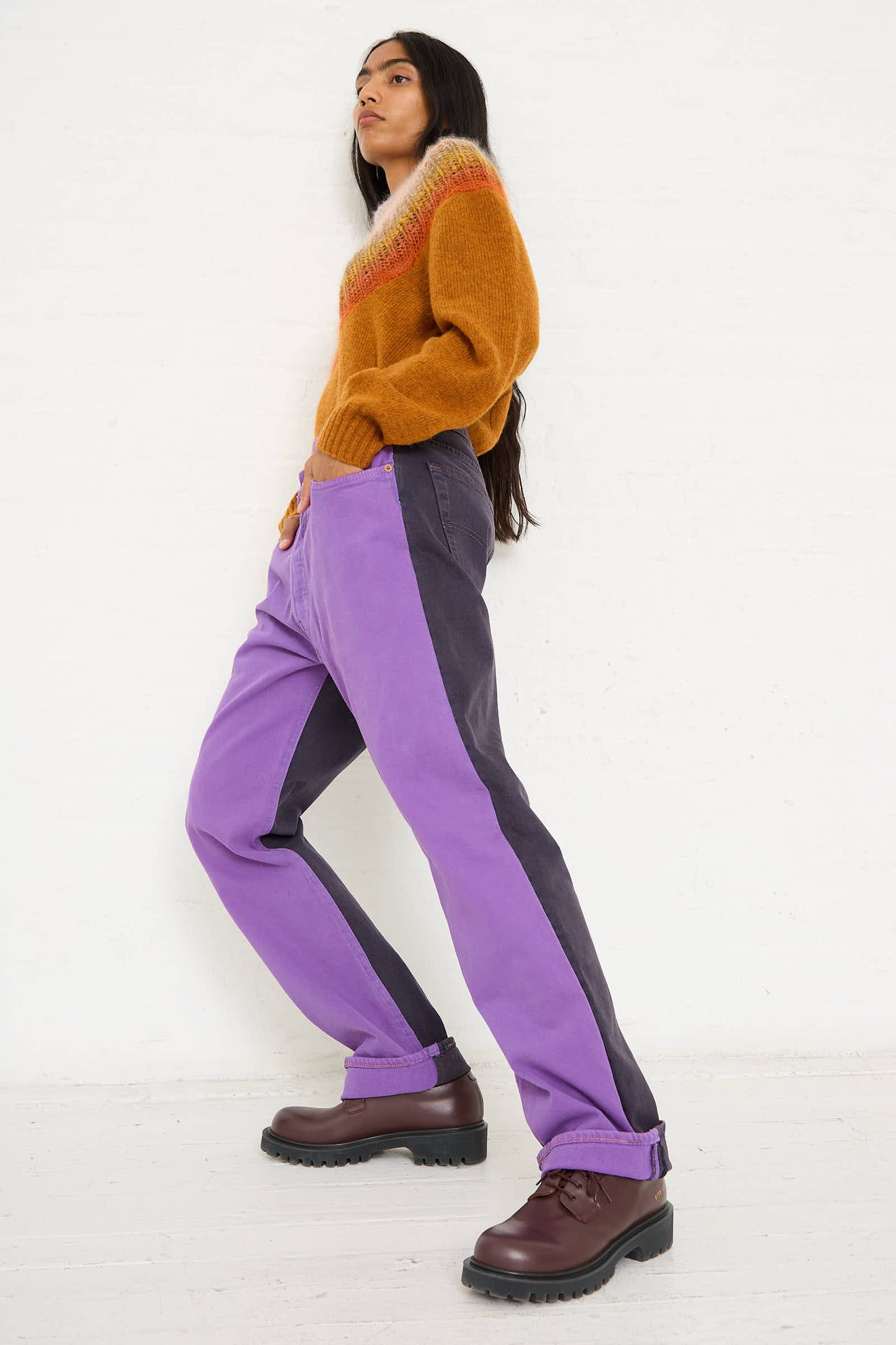 A person stands against a white wall, wearing a mustard sweater, dark brown boots, and the Bless No. 73 Jeanspleatfront in Purple/Black with a relaxed fit. They are positioned slightly sideways with hands in both pockets.