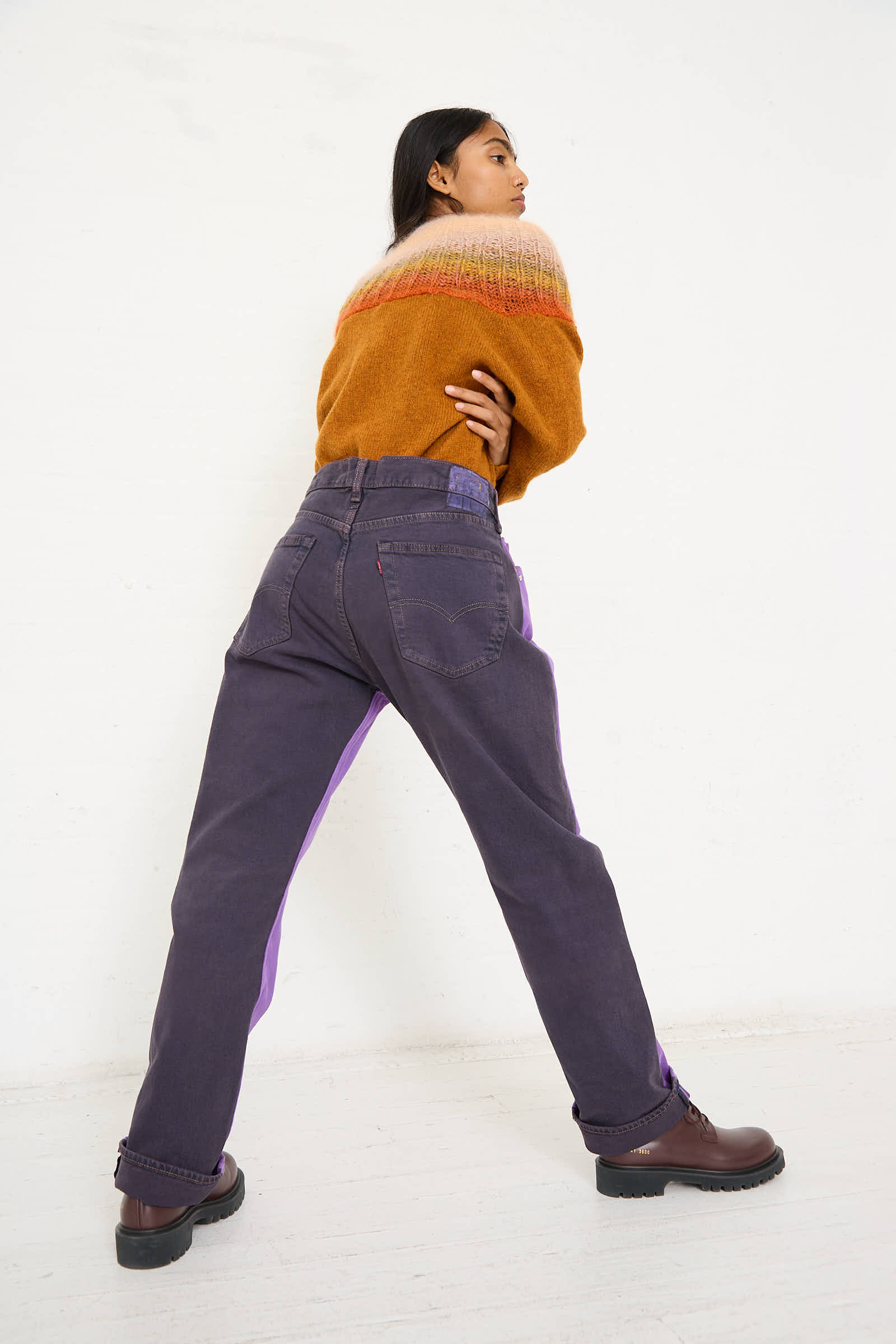 A person stands facing backward, wearing a mustard sweater with multicolored stripes at the top, No. 73 Jeanspleatfront in Purple/Black from Bless, and brown boots, against a plain white background.