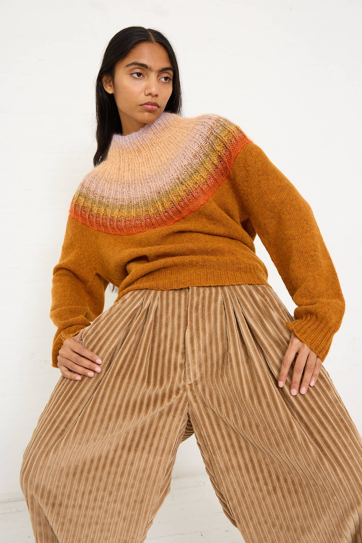 Person in a Bless No. 77 Passenpulli Chiengora Sweater in Orange with a multicolored yoke and beige striped wide-leg pants stands against a white background, leaning slightly with one hand on their hip.