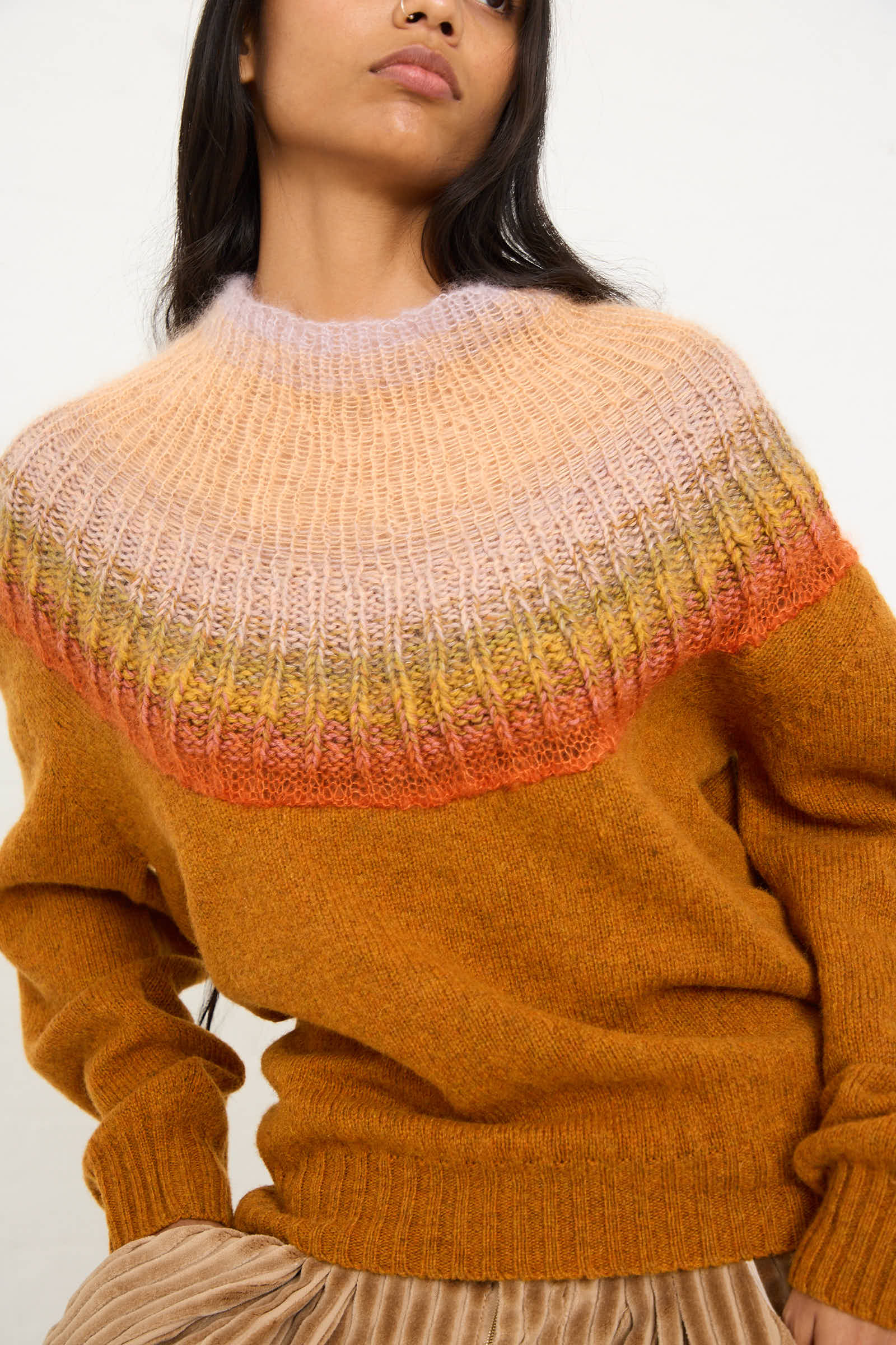 A person wearing the No. 77 Passenpulli Chiengora Sweater in Orange by Bless, featuring a hand-knitted, multicolored gradient yoke pattern and a mustard-brown body, paired with beige pleated pants.