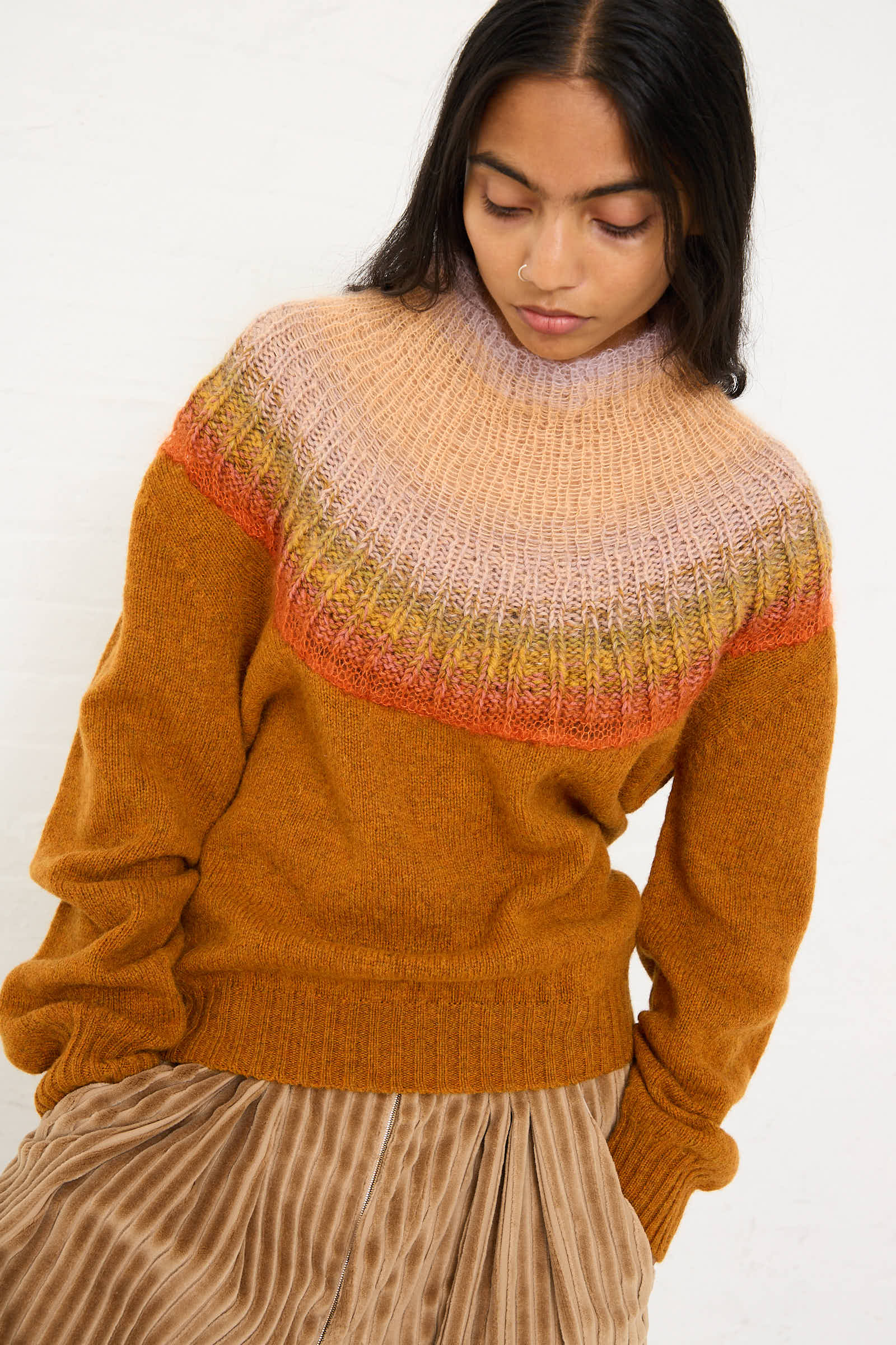 A person is wearing the cozy No. 77 Passenpulli Chiengora Sweater in Orange by Bless, featuring a hand-knitted multicolored yoke pattern, paired with a pleated skirt, and looking down.