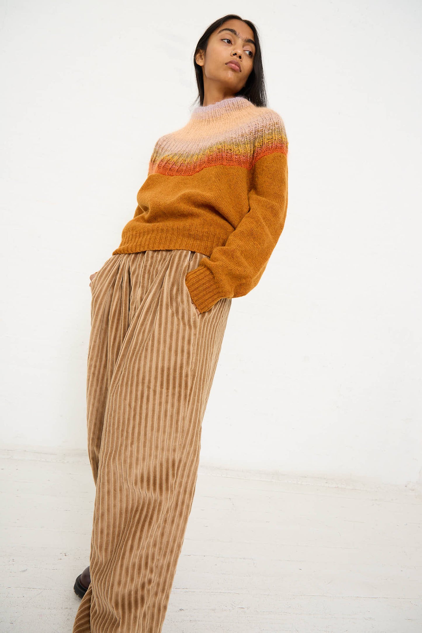 Person standing against a plain background, wearing the No. 77 Passenpulli Chiengora Sweater in Orange by Bless and beige striped wide-leg pants, with hands in their pockets.