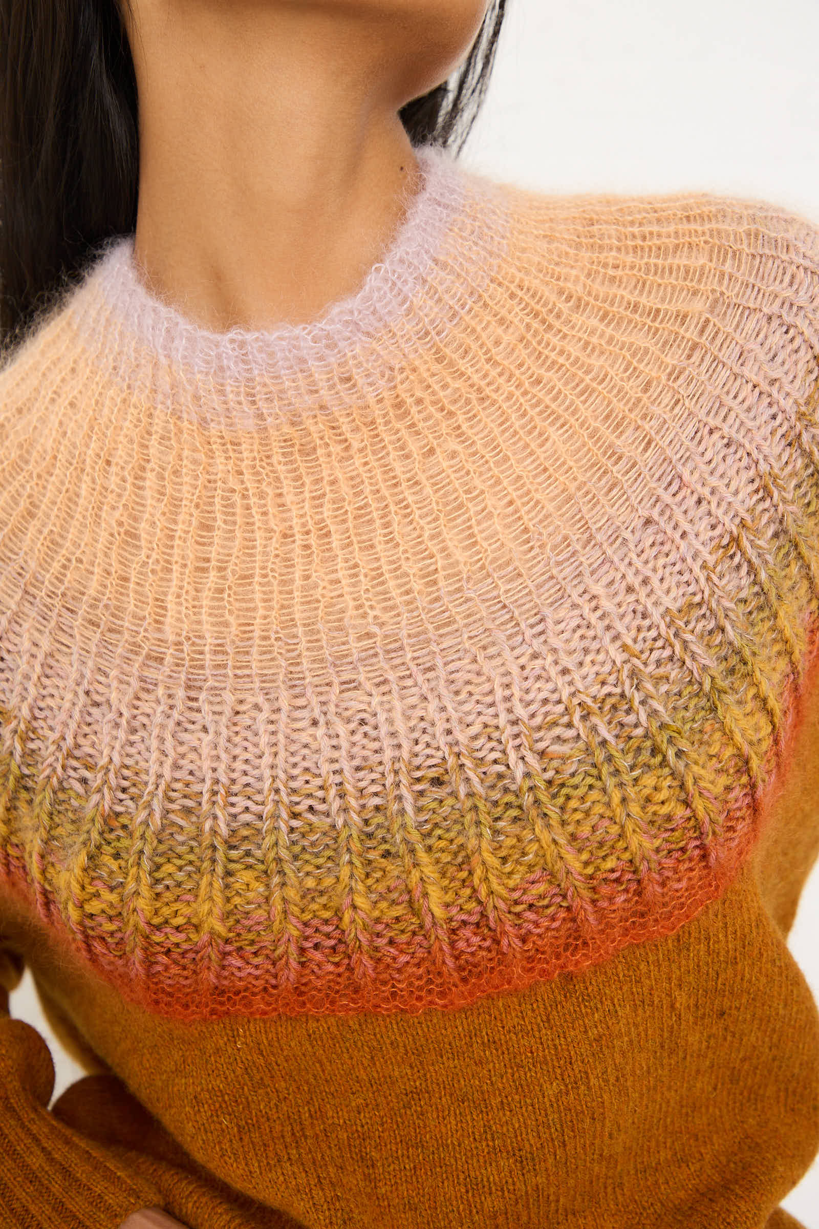 A close-up of a person wearing the No. 77 Passenpulli Chiengora Sweater in Orange by Bless, showcasing the gradient pattern in shades of orange, yellow, and purple. The image highlights the upper chest and collar of this luxurious wool/cashmere/mohair blend garment.