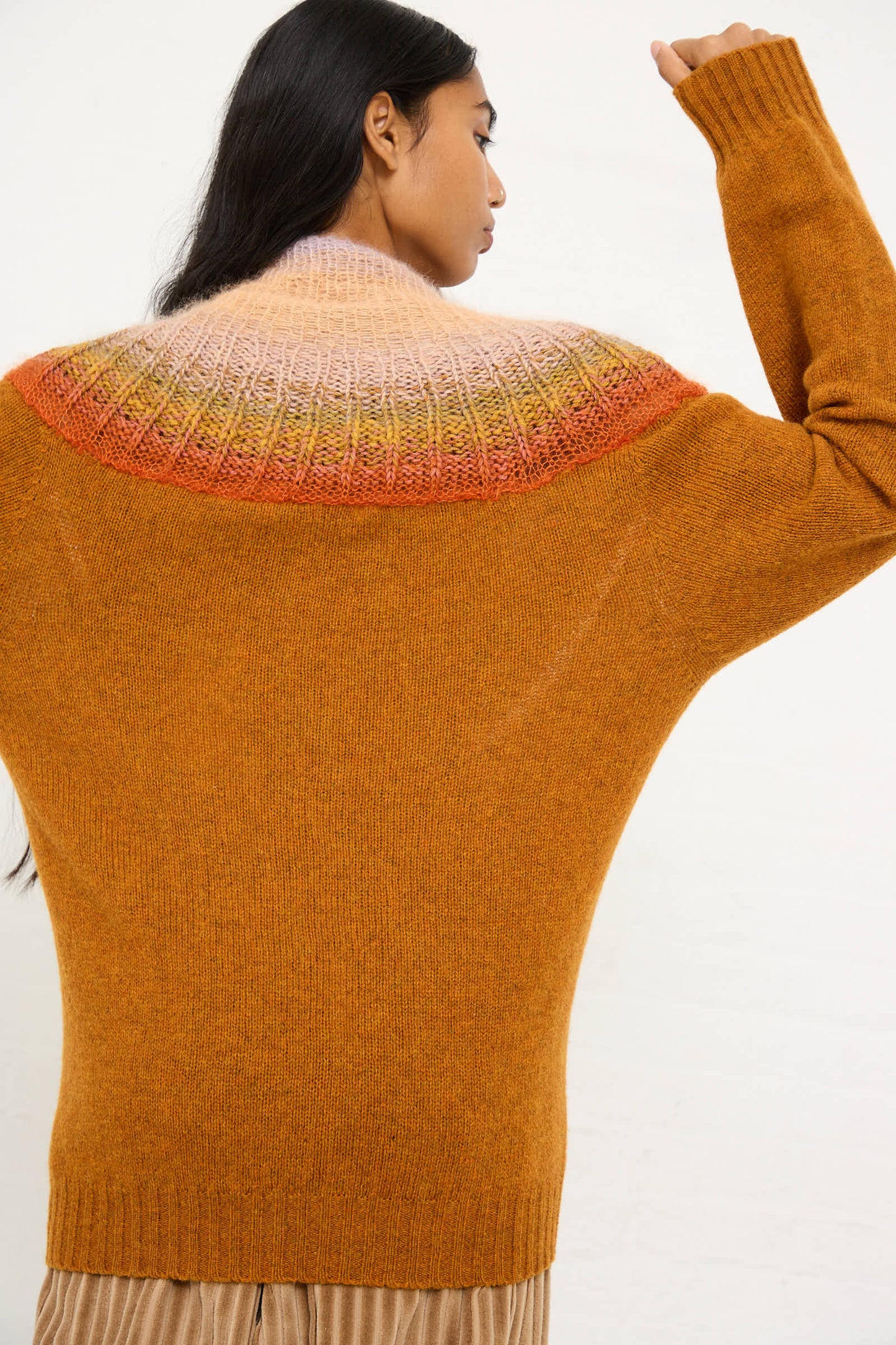 A person with long dark hair is shown from behind, wearing the No. 77 Passenpulli Chiengora Sweater in Orange by Bless, a rust-colored, hand-knitted piece crafted from a luxurious blend of wool, cashmere, and mohair. The sweater features a stunning gradient yoke design in pink, orange, and yellow shades.