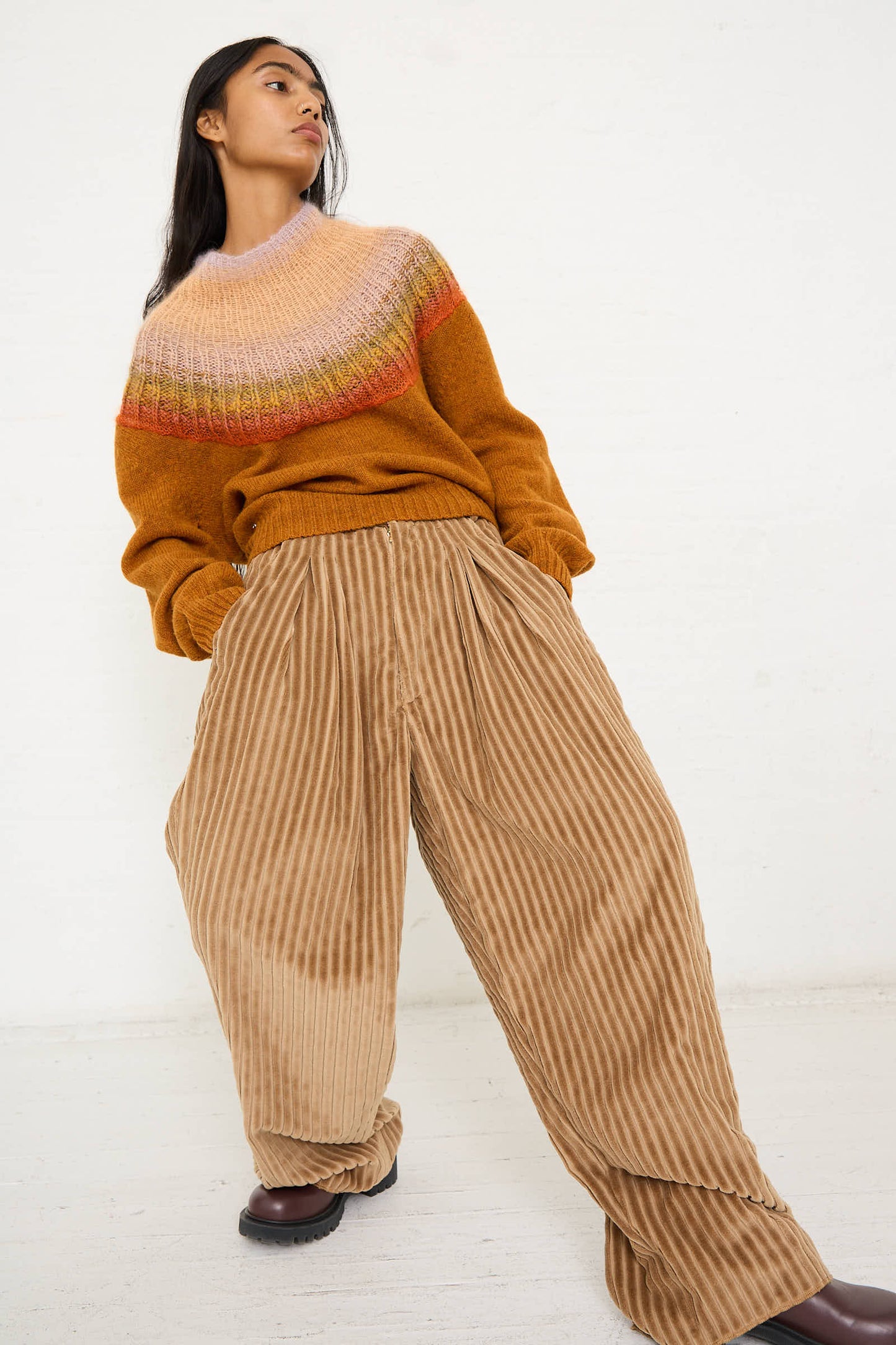 Person wearing an orange and pink sweater, Bless's No. 78 Ultrawidepleated Pant in Jumbo Beige Cord, and brown shoes poses against a plain white background.