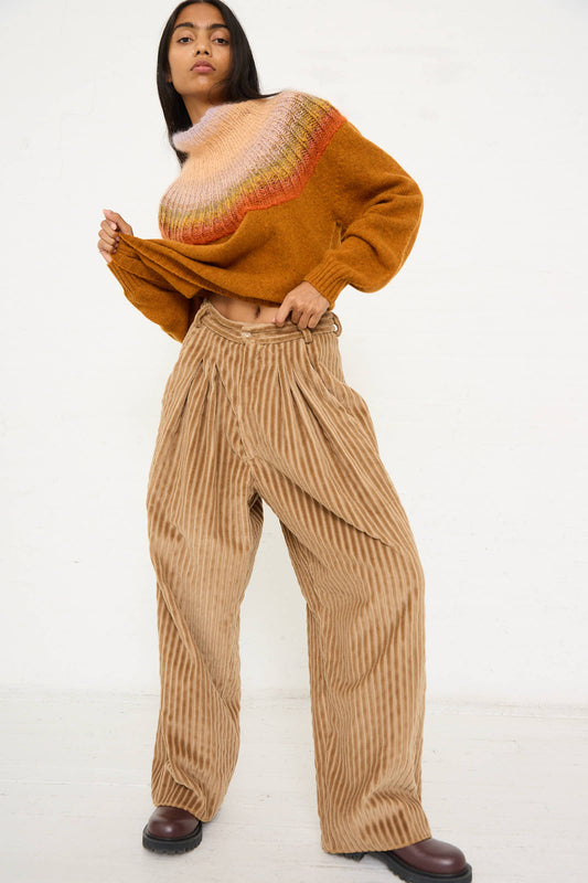 A person stands against a white background wearing a multicolored sweater and Bless's No. 78 Ultrawidepleated Pant in Jumbo Beige Cord, paired with dark brown shoes. They have a relaxed pose with one hand on their sweater hem.