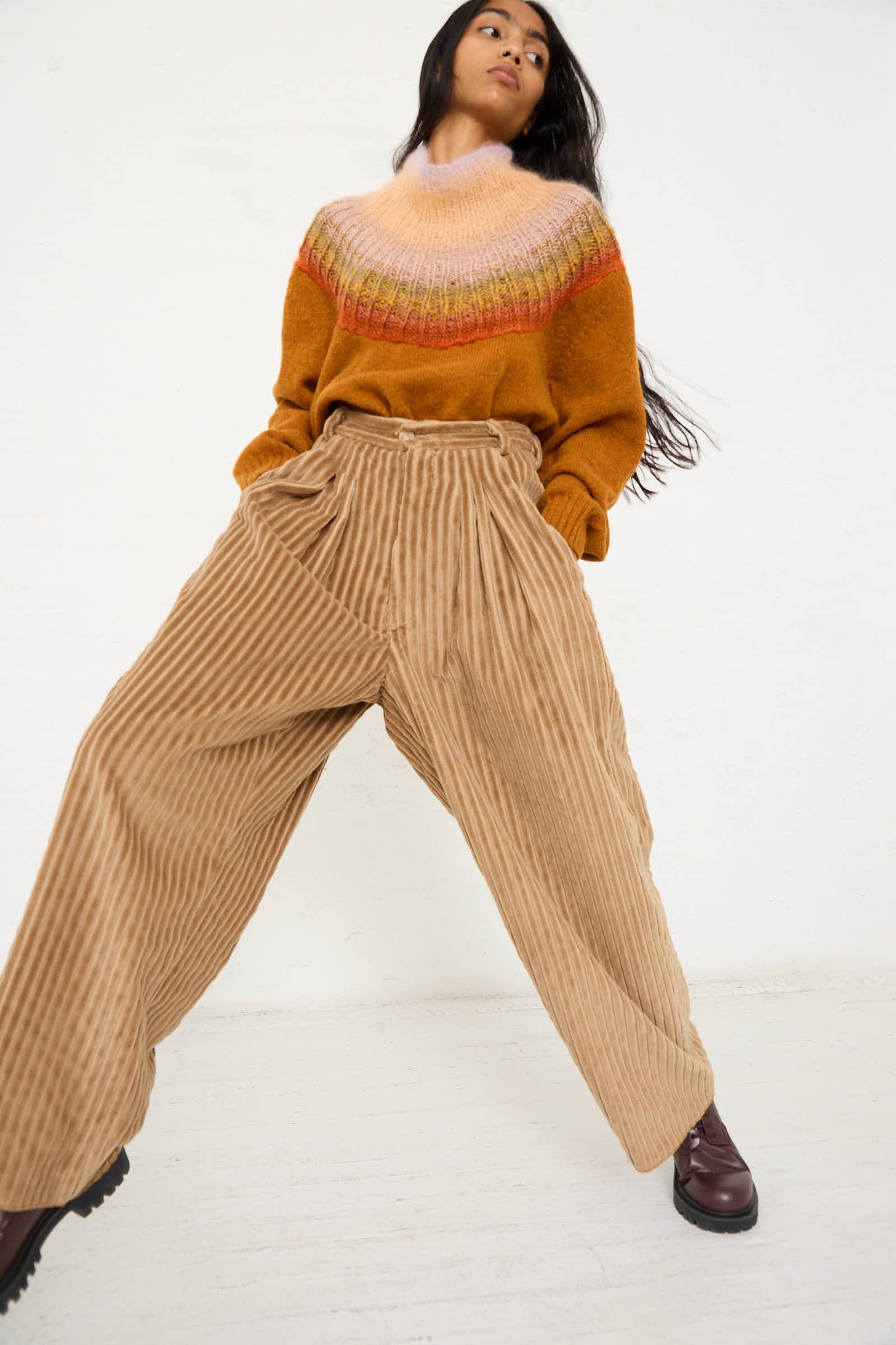 Person wearing a colorful, gradient sweater and the No. 78 Ultrawidepleated Pant in Jumbo Beige Cord by Bless stands with a slight lean against a white background.