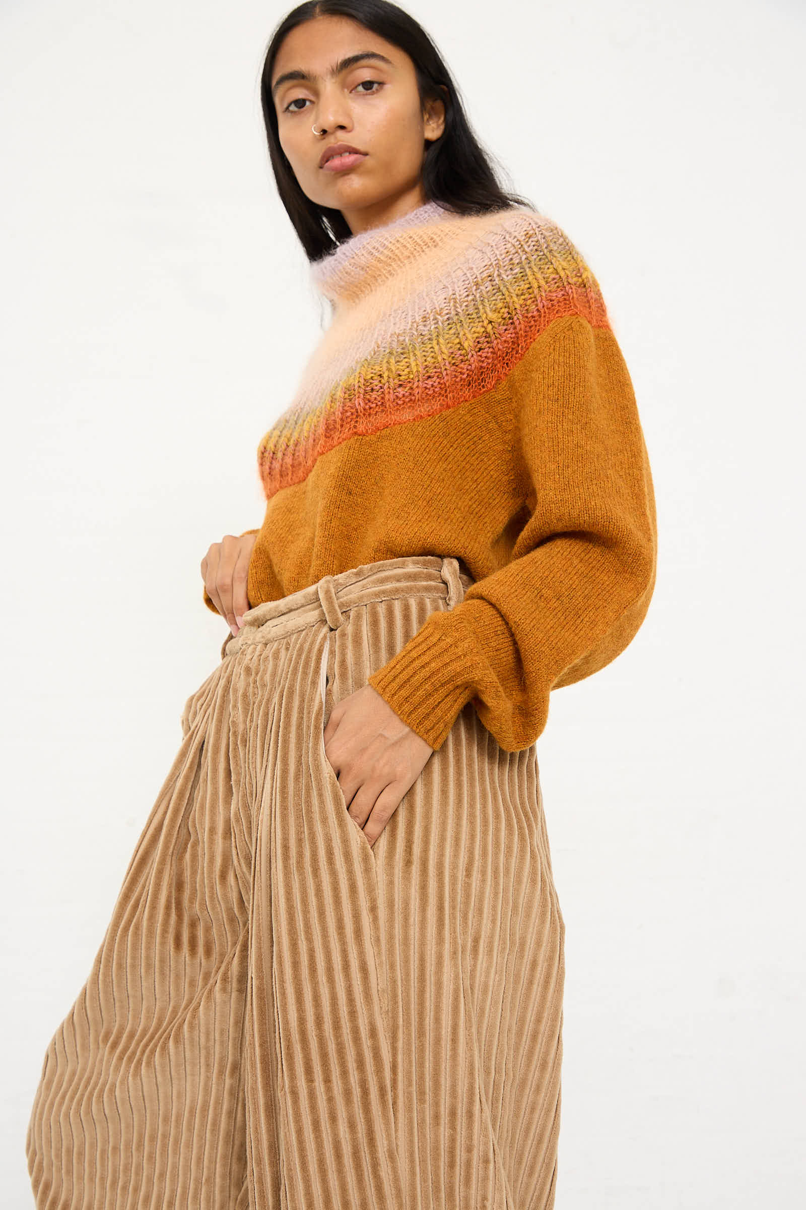 Person in a multicolored sweater and Bless No. 78 Ultrawidepleated Pant in Jumbo Beige Cord, with hand in pocket, posing against a plain background.
