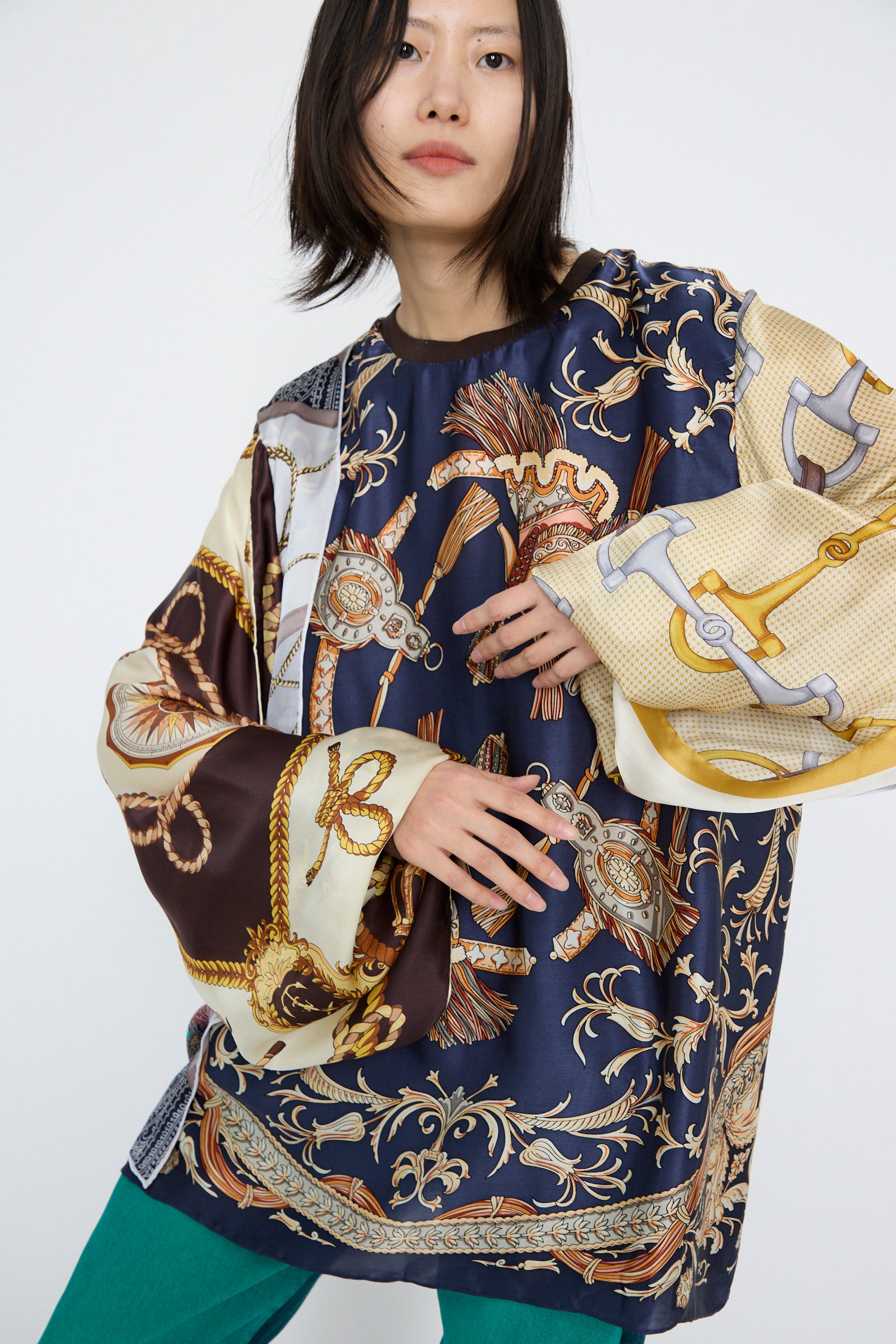 A person in an oversized Bless Windy Silkscarves Top, featuring green, brown, and yellow prints reminiscent of vintage scarves with nautical and floral designs, looks at the camera against a neutral background.