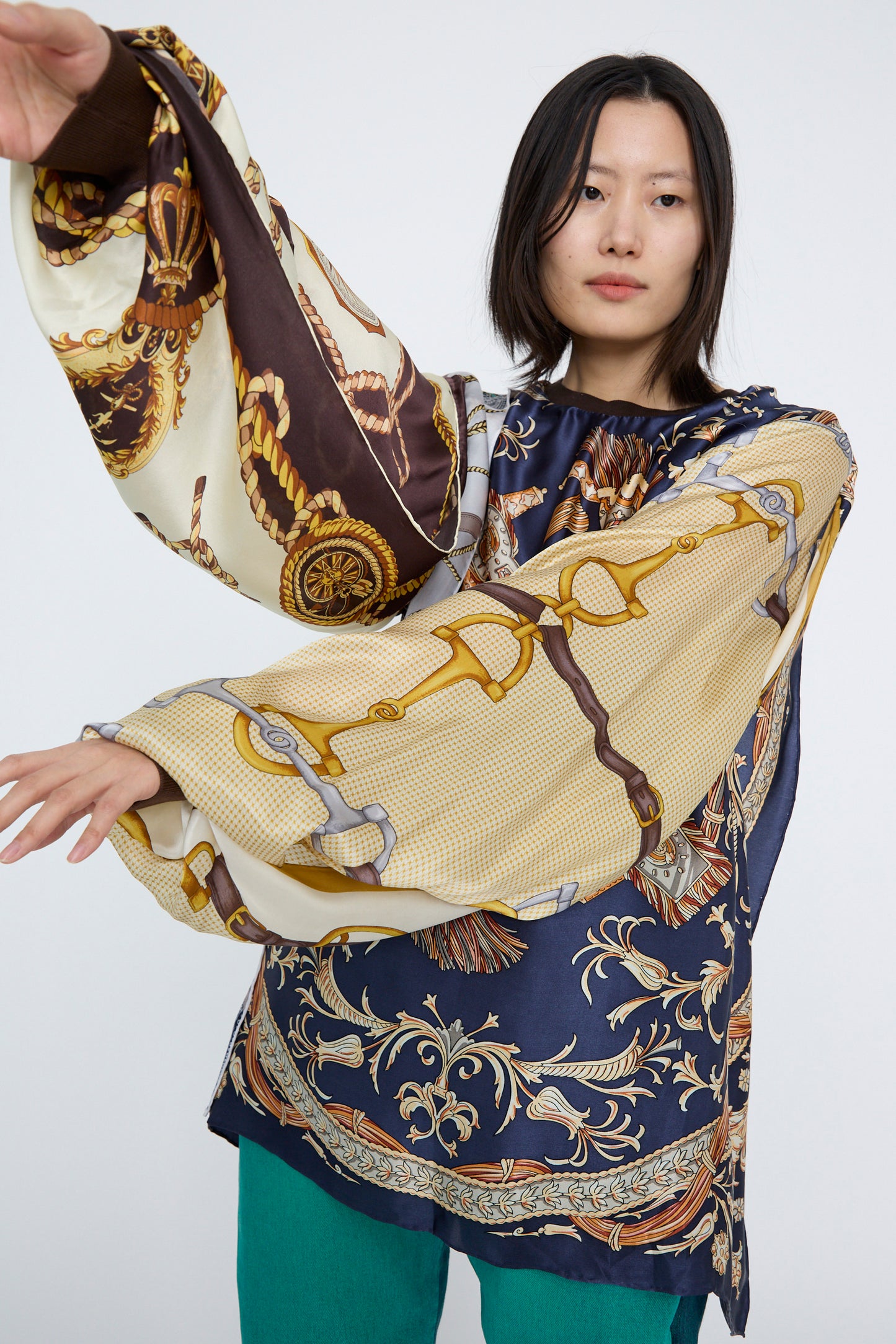 A person with dark hair extends their arms outward against a plain background, wearing an oversized Bless Windy Silkscarves Top in Green Brown Yellow Prints I featuring colorful abstract patterns and wide sleeves, paired with teal pants.