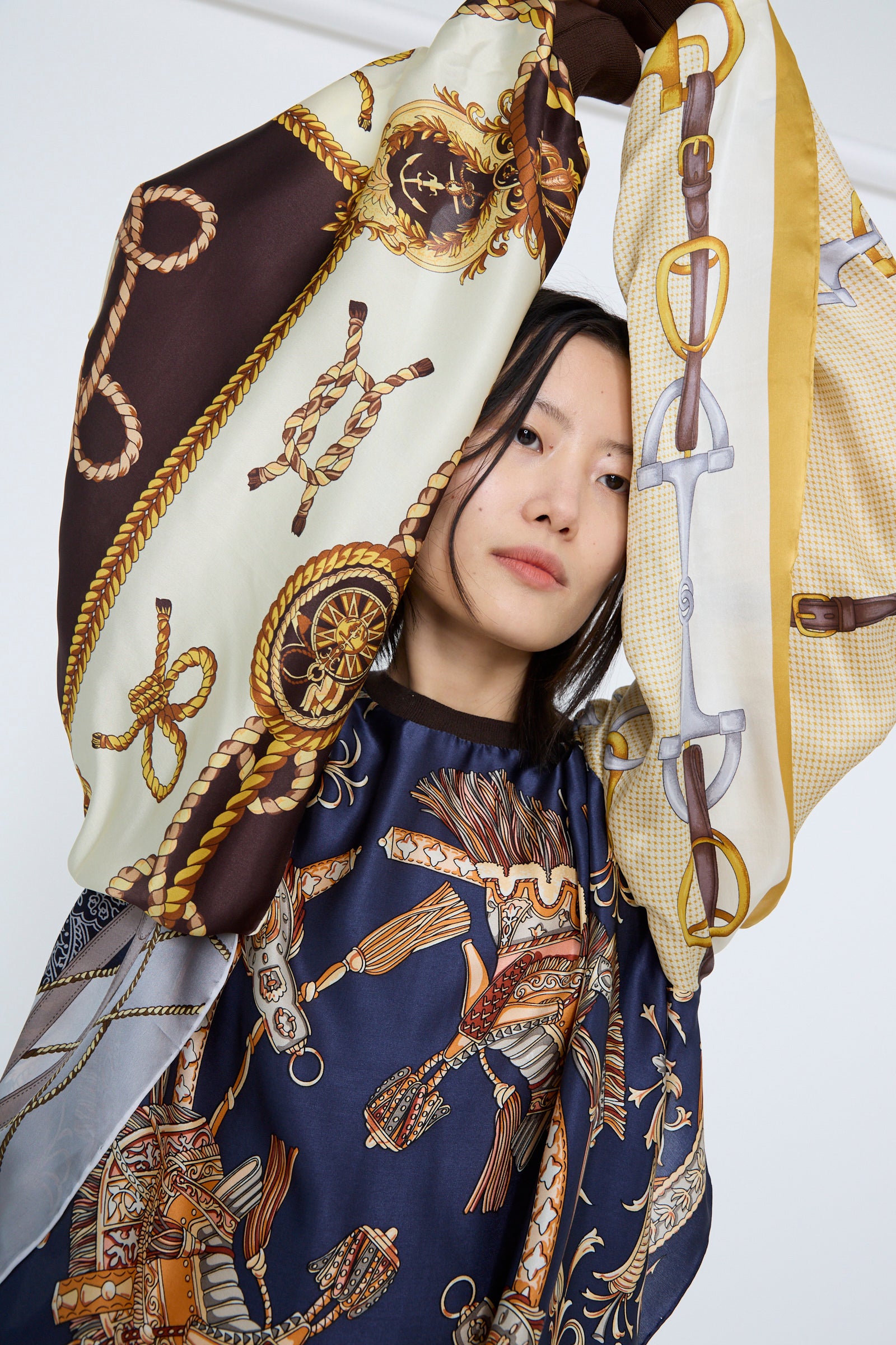 A woman wears the Windy Silkscarves Top in Green Brown Yellow Prints I by Bless, featuring vintage equestrian and nautical designs in brown, yellow, and navy blue, her arms raised against a white backdrop. 