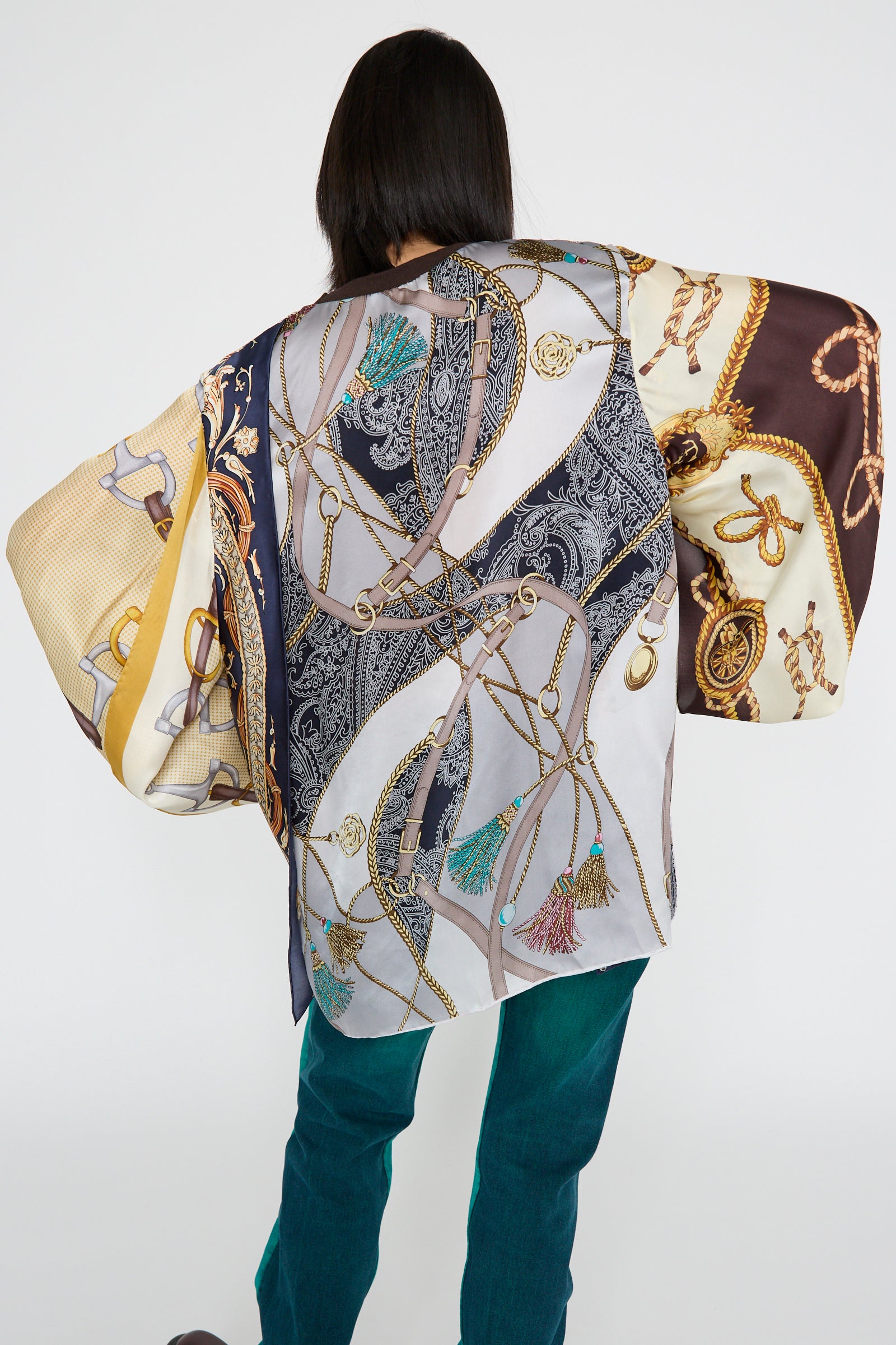 A person models the "Windy Silkscarves Top in Green Brown Yellow Prints I" by Bless, a 100% silk oversized top with colorful, vintage scarf-inspired patterns featuring intricate rope and chain designs, against a plain backdrop.