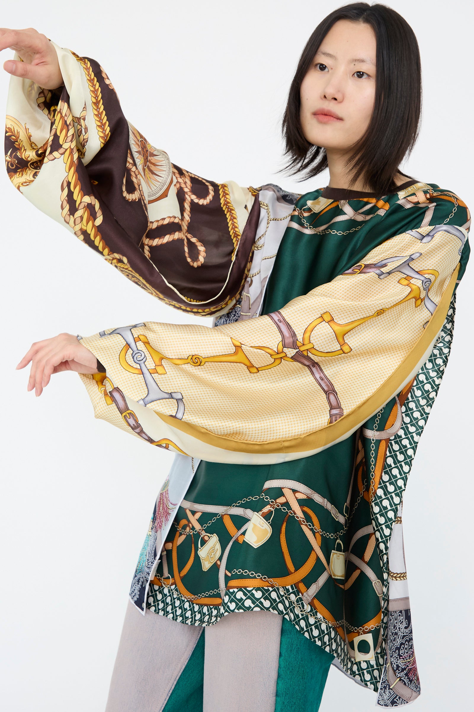 A person wearing the Windy Silkscarves Top in Green Brown Yellow Prints II by Bless, featuring intricate patterns and long flowing sleeves, stands against a plain white background with arms raised. 