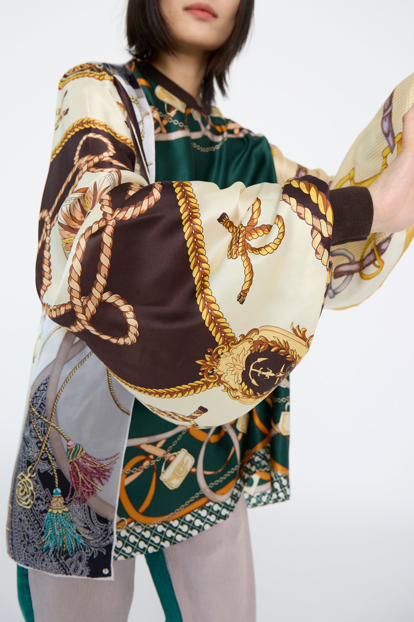 A person wearing the Windy Silkscarves Top in Green Brown Yellow Prints II by Bless, featuring oversized rope and chain designs reminiscent of vintage scarves, against a white backdrop.