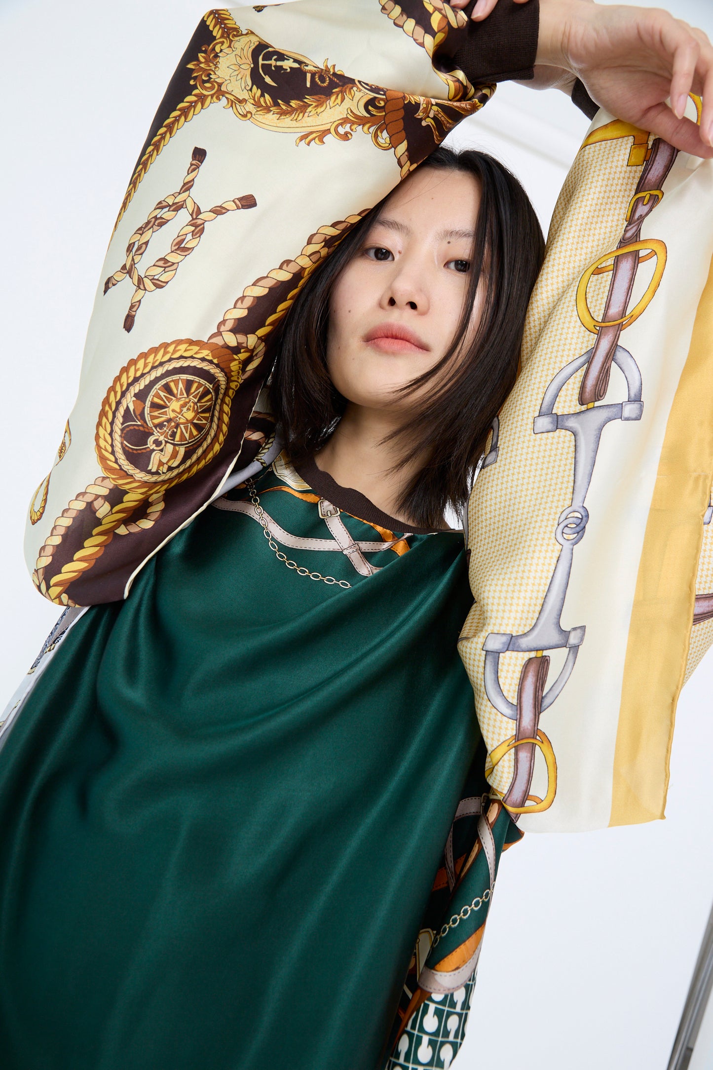 A person wearing Bless's Windy Silkscarves Top in Green Brown Yellow Prints II, featuring oversized styles and sleeves with vintage scarf-like designs of ropes and chains. Their arms are raised above their head.