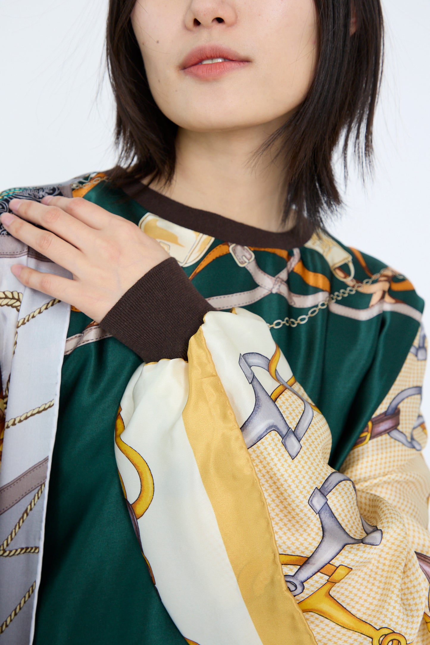 A model wearing the Windy Silkscarves Top in Green Brown Yellow Prints II by Bless rests a hand on their shoulder. The oversized shirt features colorful patterns and chain designs, with 100% silk fabric draping like vintage scarves.