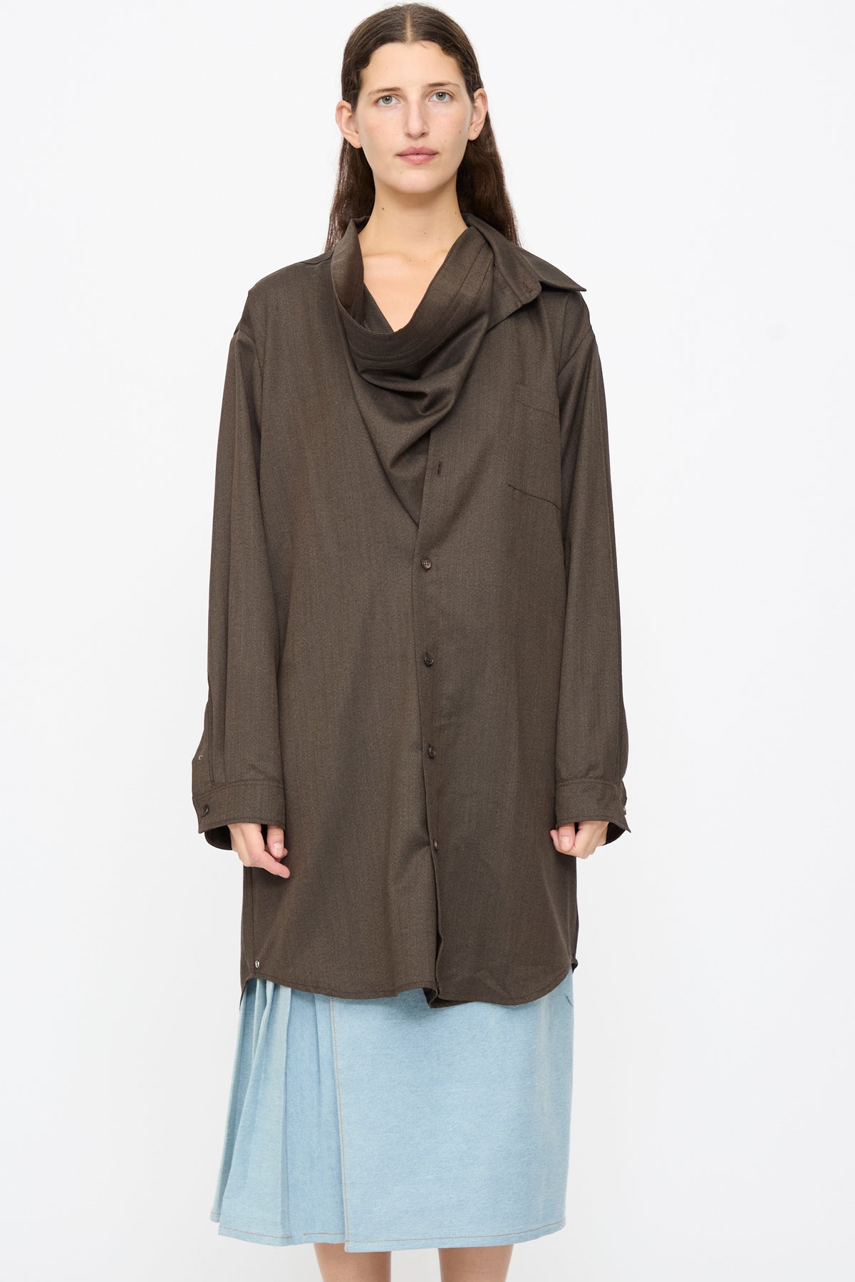 A person wears a Bless Wool No. 82 Neckerchiefdress in Brown Melange, an oversized, brown wool button-down shirt with an asymmetrical scarf collar over a light blue skirt.