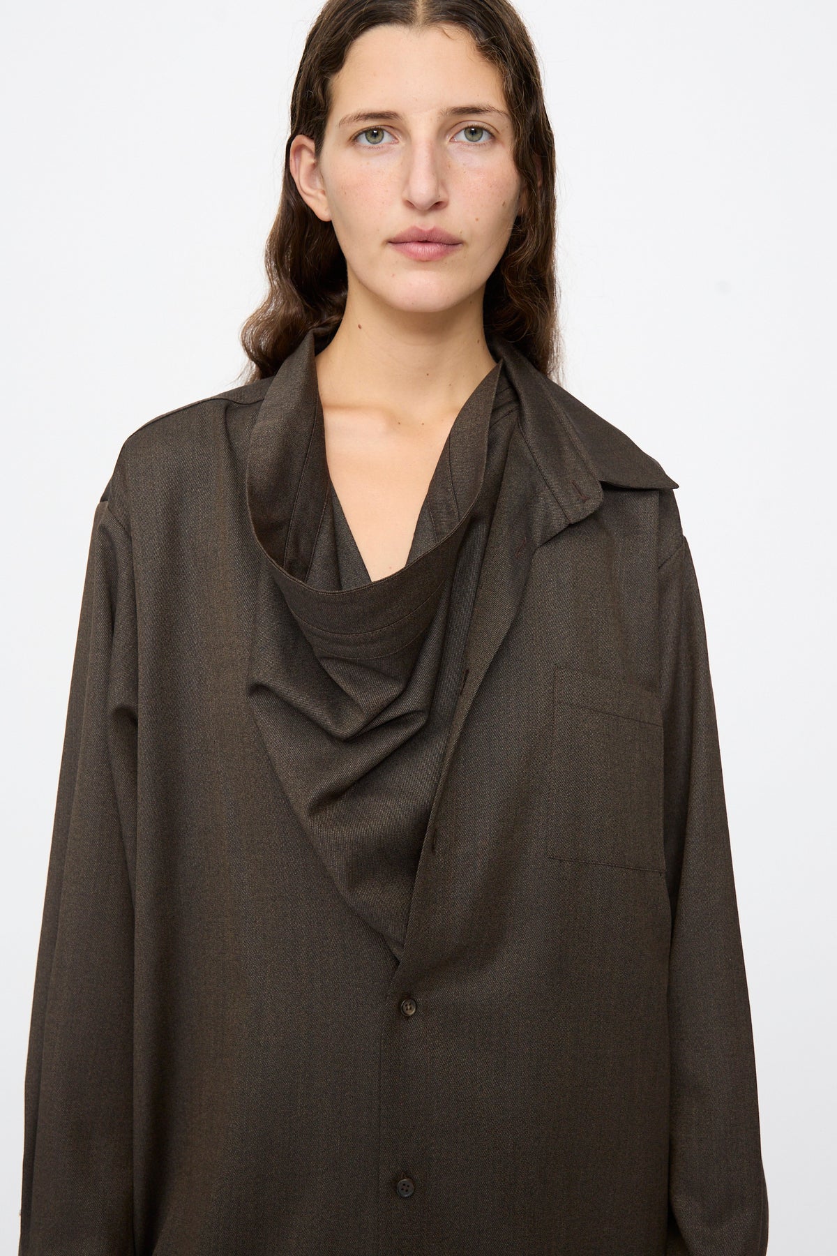A woman wears a Bless Wool No. 82 Neckerchiefdress in brown melange, an oversized button-up shirt-dress with an asymmetric, draped cowl neckline.
