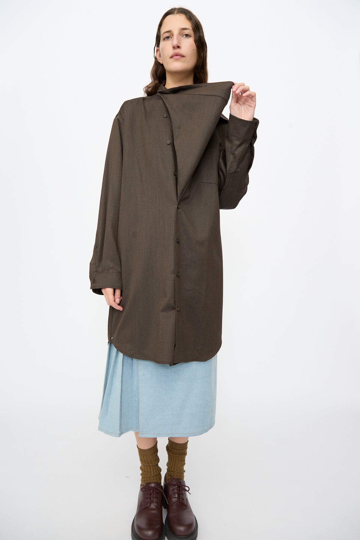 A person wears a Bless Wool No. 82 Neckerchiefdress in Brown Melange with an asymmetrical scarf collar over a light blue denim midi skirt, brown socks, and dark brown leather shoes.