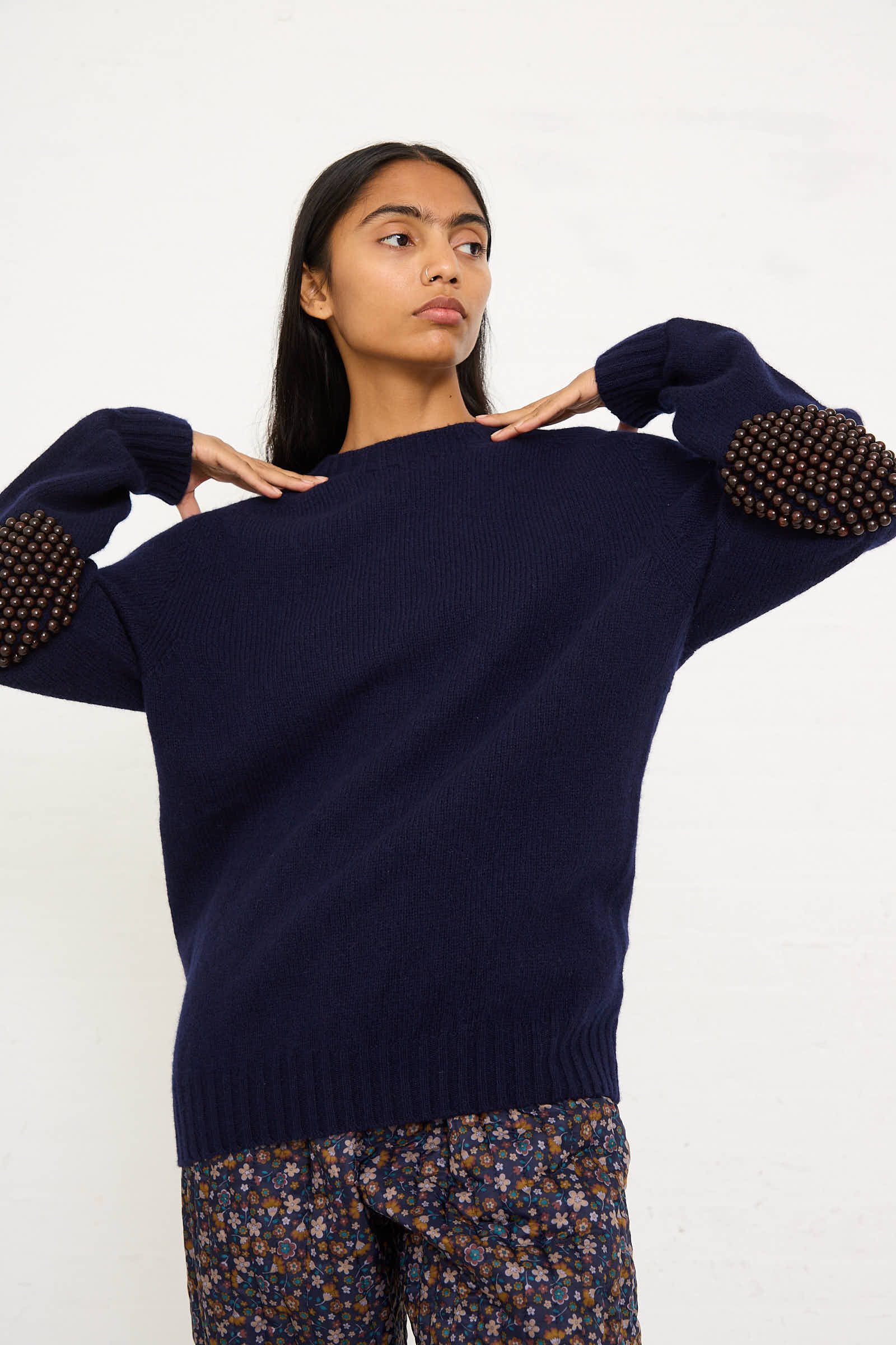 A person wearing the Bless Wool No. 75 Pearlpad Sweater in Navy with Wooden Pearls and patterned pants stands against a plain background.