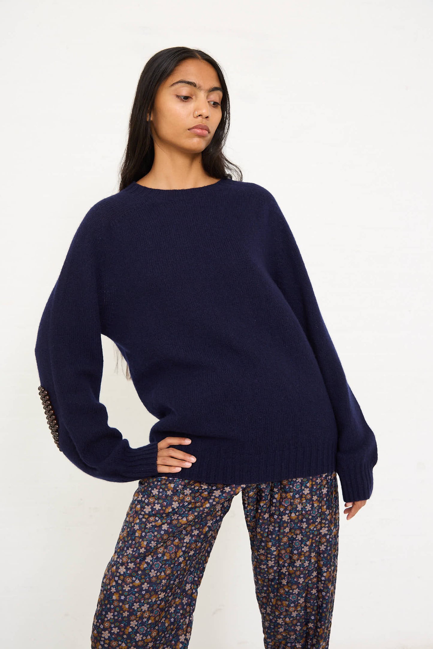 A person stands against a white background, wearing the loose-fitting navy blue Wool No. 75 Pearlpad Sweater in Navy with Wooden Pearls by Bless and patterned pants. The sweater, featuring a button detail on one sleeve and beaded elbow patches, complements their long, dark hair as they gaze to the side.