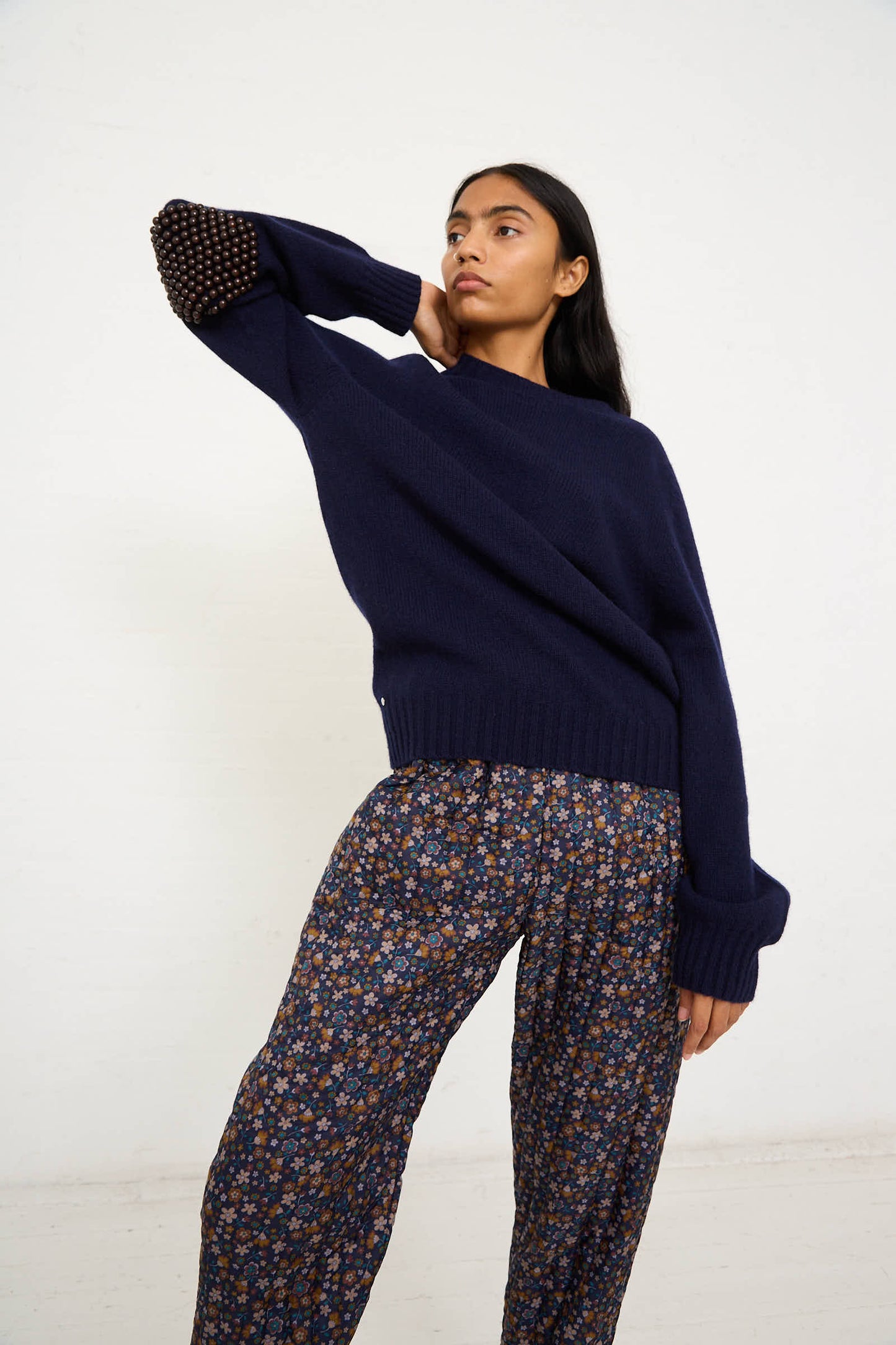 A person posing against a plain background, wearing the Bless Wool No. 75 Pearlpad Sweater in Navy with Wooden Pearls and patterned pants, with one hand behind their head.