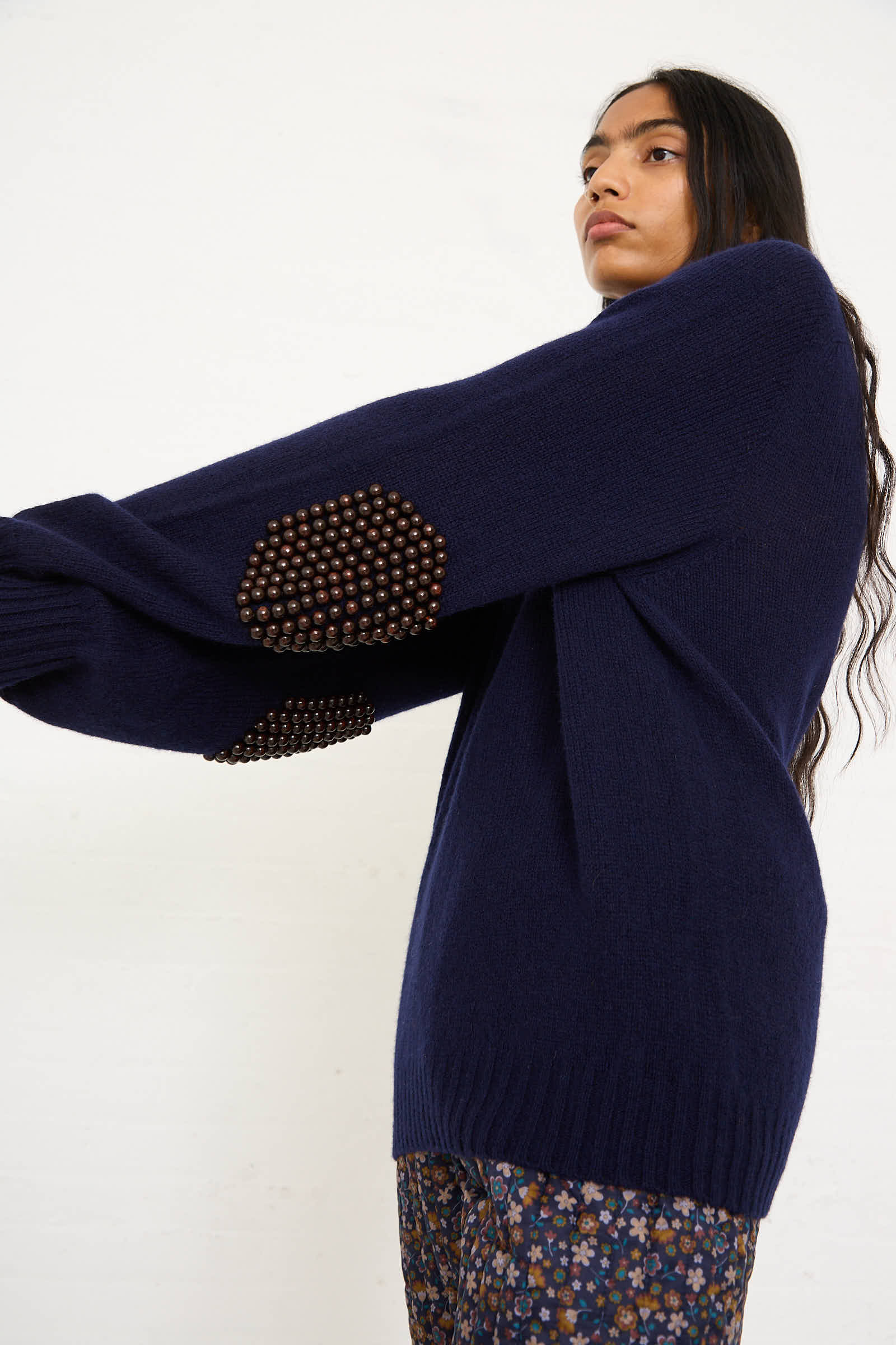 A person wearing the Bless Wool No. 75 Pearlpad Sweater in Navy with Wooden Pearls, standing against a plain white background and wearing floral-patterned pants.