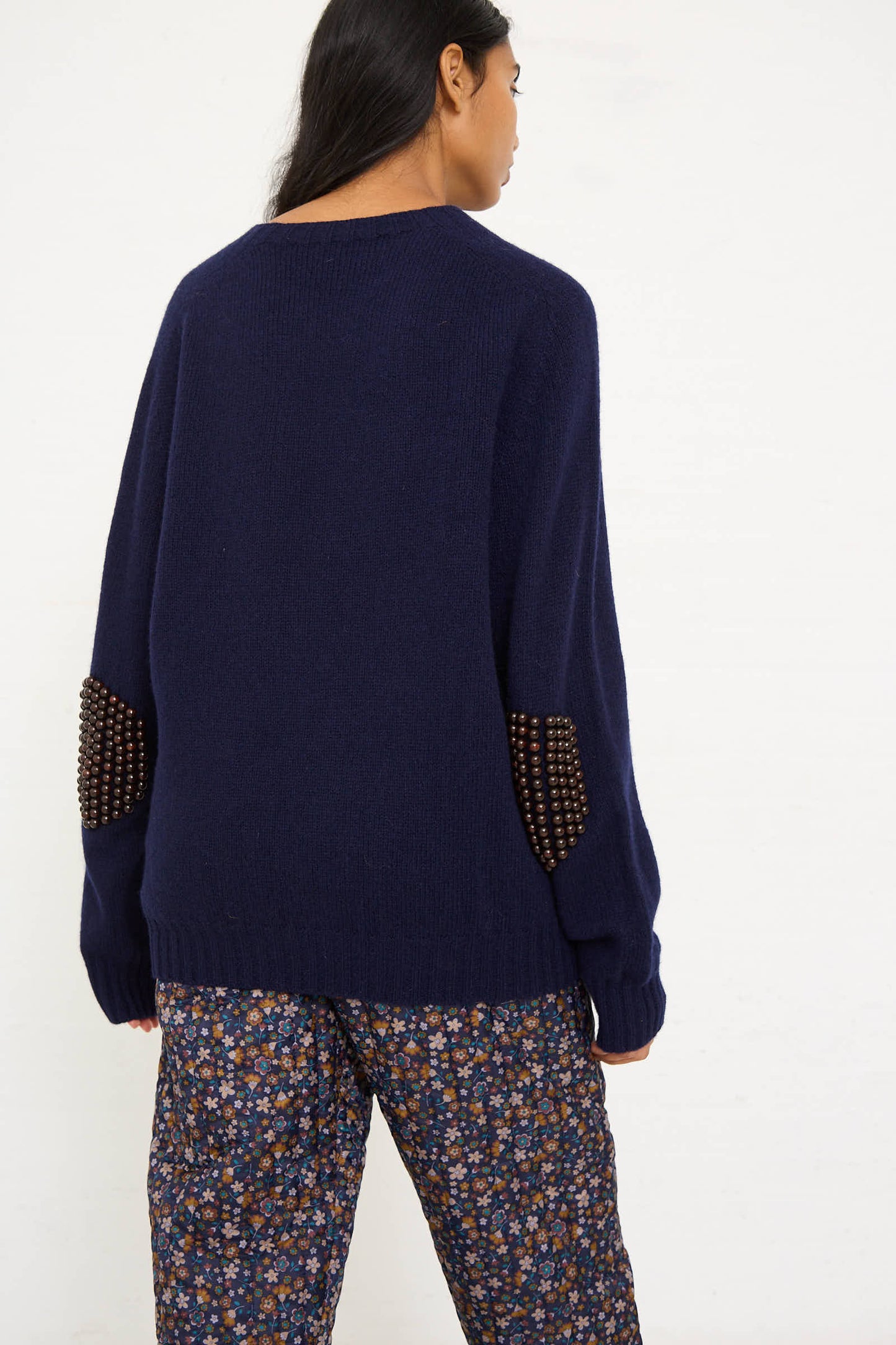 A person wearing the Bless Wool No. 75 Pearlpad Sweater in Navy with Wooden Pearls and floral patterned pants, facing away from the camera.