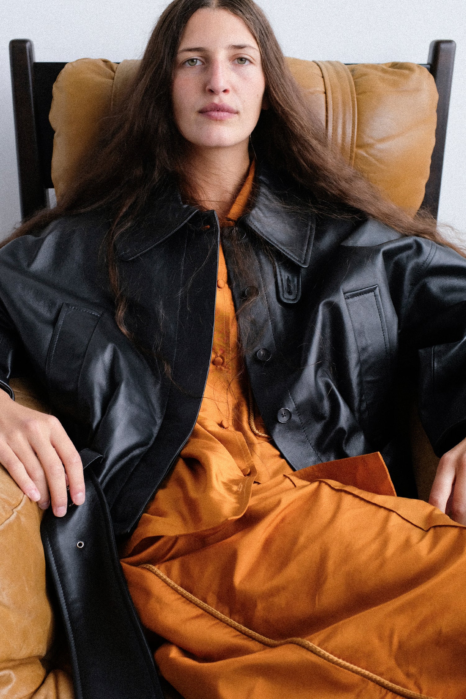 A woman with long dark hair sits in a tan leather armchair, wearing Cawley's Silk Dupion August Shirt in Sienna, Silk Dupion Luna Trouser with Trim in Sienna, and the Nappa Leather Nara Jacket in Black.