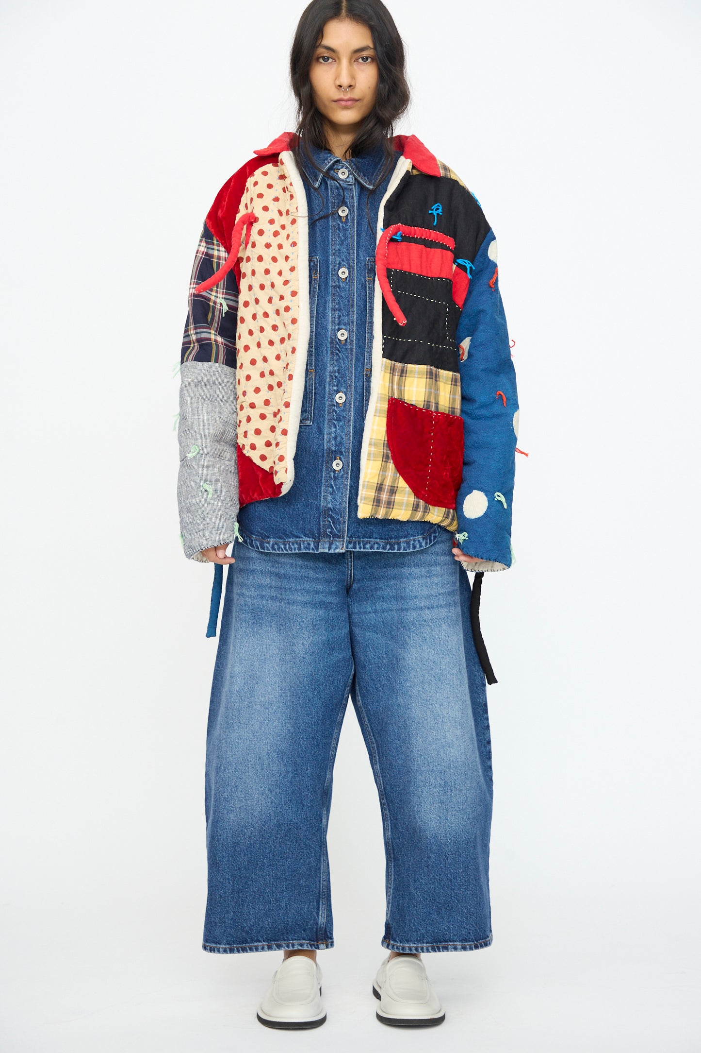 A person stands facing forward wearing the CMC Polka Assembly Coat, a hand-quilted patchwork piece made in New York, along with a denim shirt, wide-leg jeans, and white shoes against a plain white background.