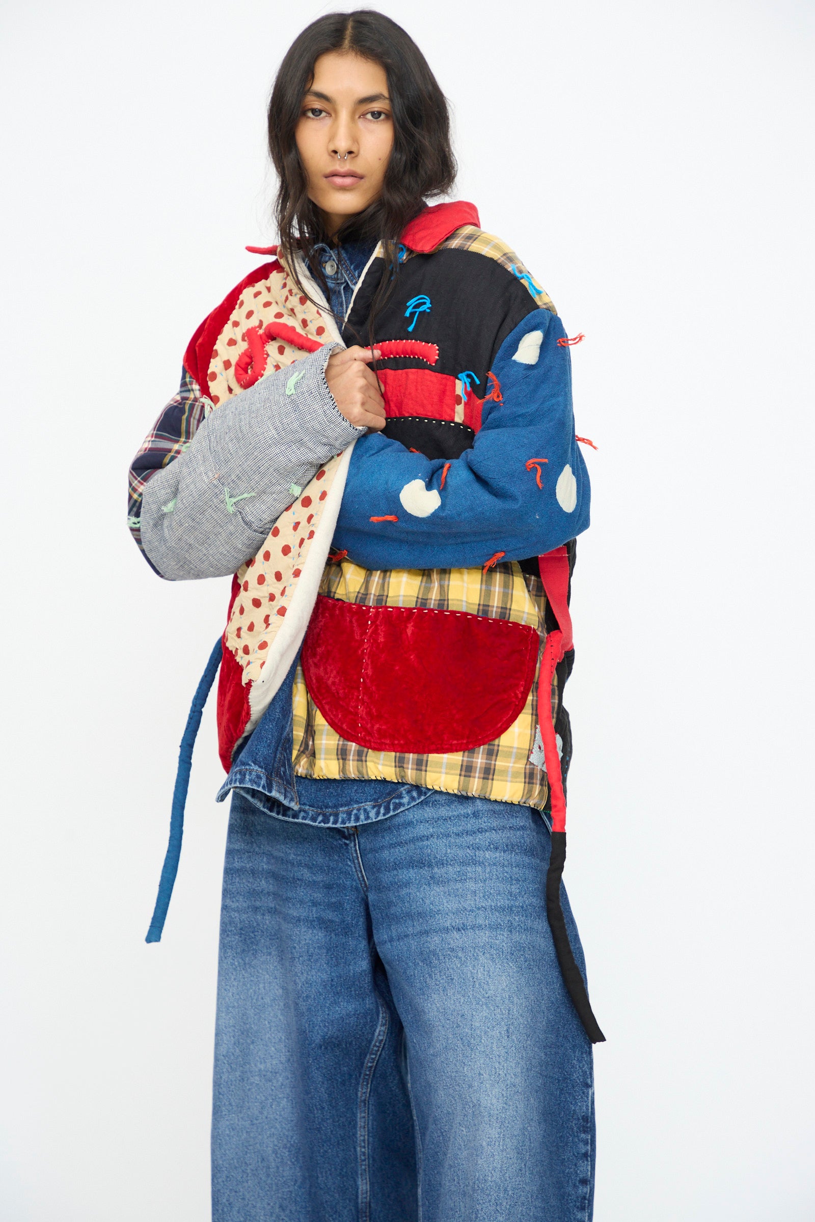 A model wears the Polka Assembly Coat by CMC, a hand-quilted patchwork piece crafted in New York from mixed fabrics and patterns, styled with wide-leg blue jeans against a plain white background.