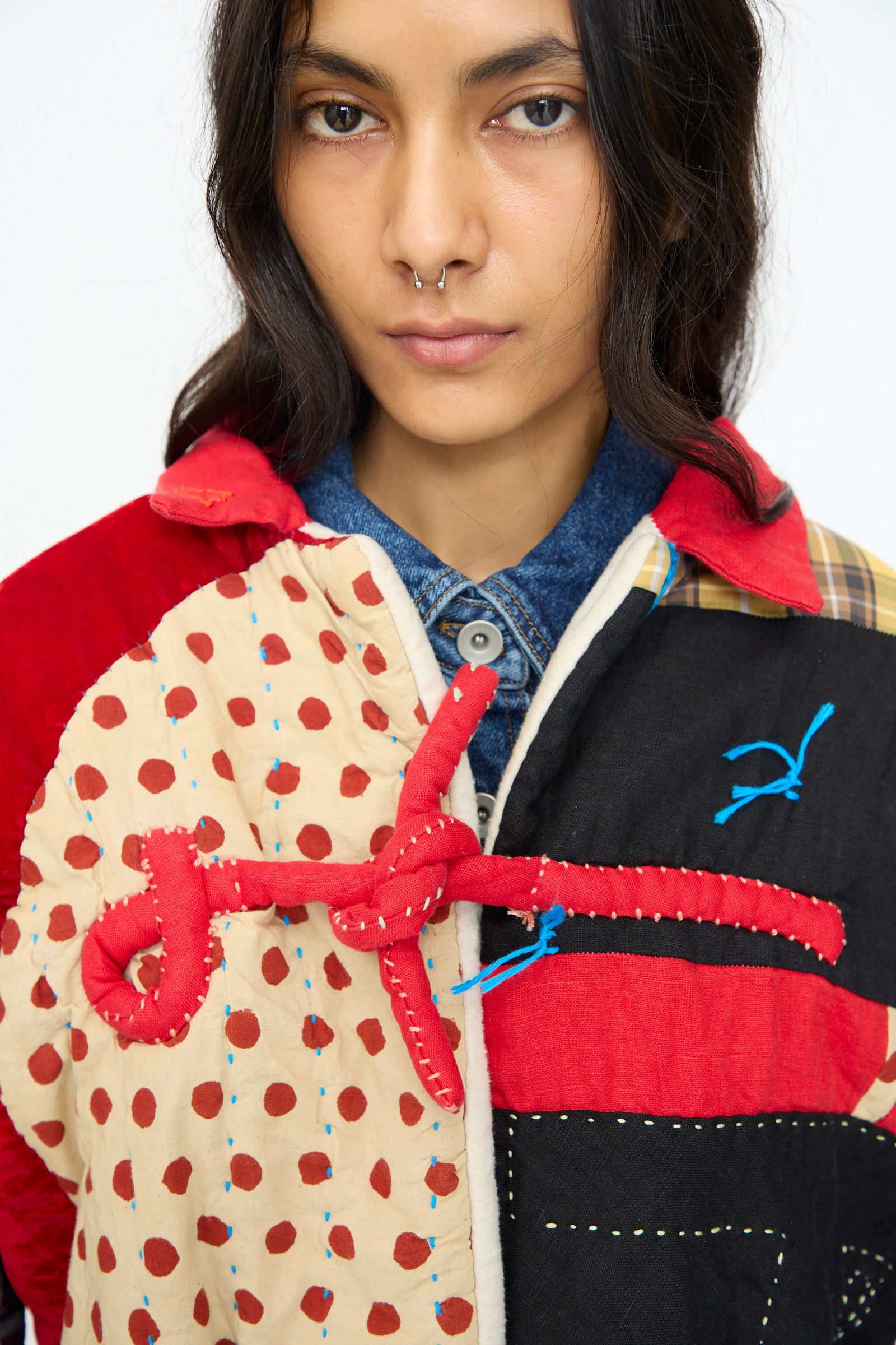 A person with long dark hair and a nose ring wears the CMC Polka Assembly Coat, a hand-quilted patchwork piece made in New York with red and beige polka dots plus blue stitching accents.