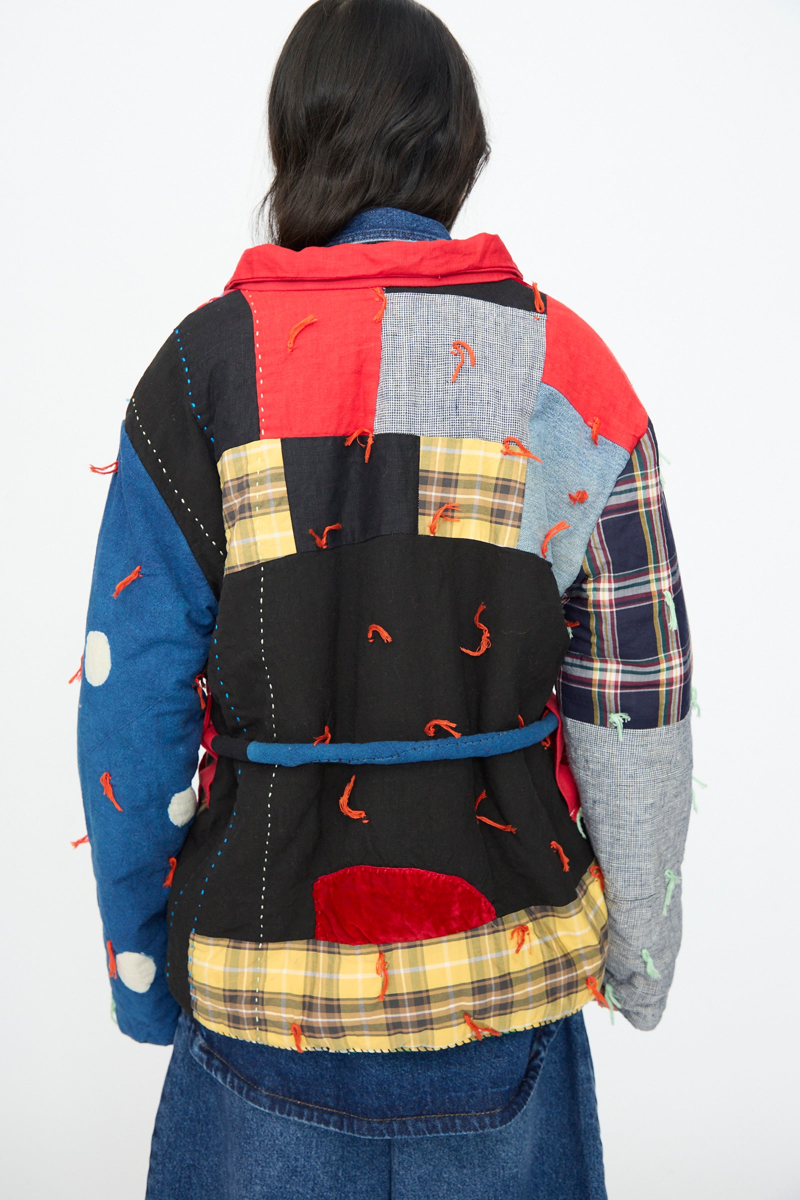 A person wears the CMC Polka Assembly Coat—a colorful, hand-quilted 100% cotton coat with mixed patterns and red stitching—paired with a denim skirt, photographed from behind. Thoughtfully made in New York.