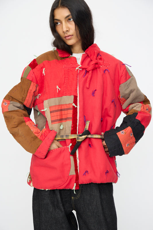 A person wearing the Red Assembly Coat II by CMC—a hand-quilted patchwork coat with mixed textures and patterns, belted at the waist—pairs it with dark jeans against a plain background. This unique piece is made in New York.