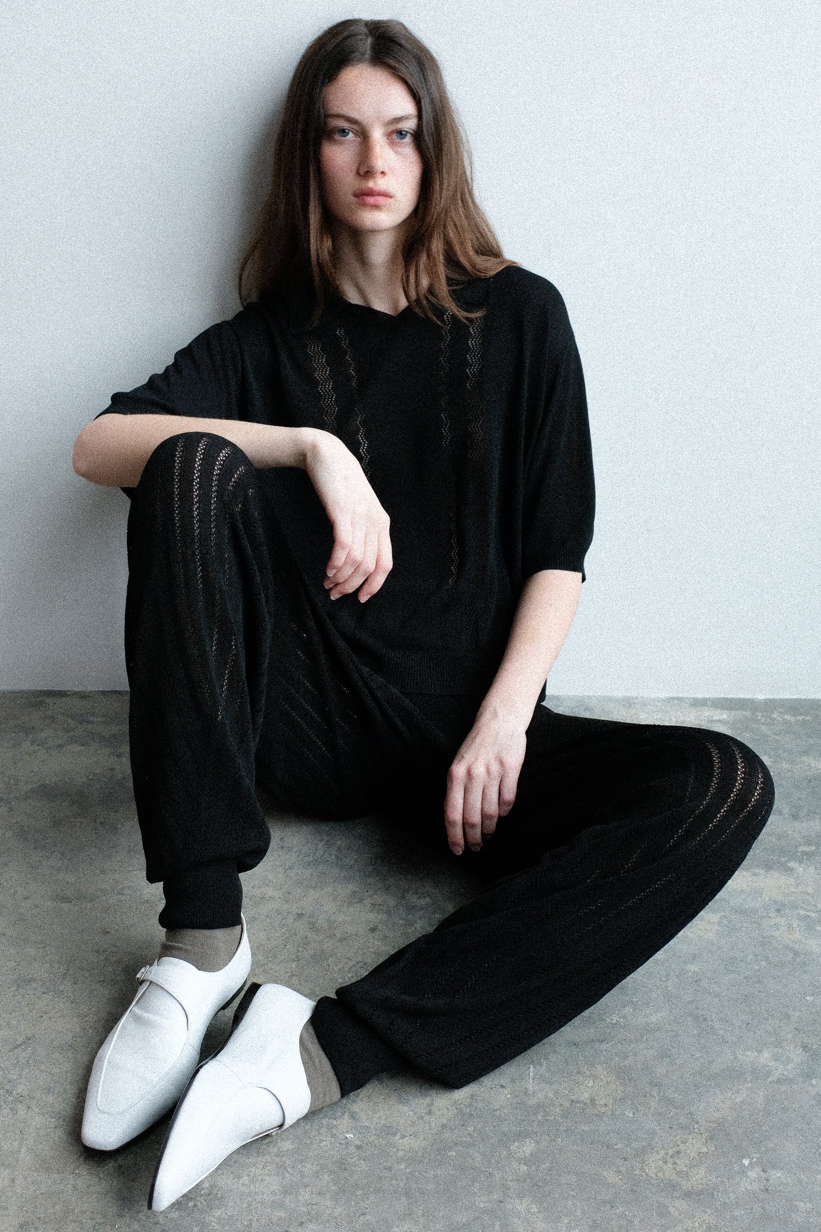 A woman sitting on a concrete floor wearing Cordera's Lace Knit Polo in Black with the Lace Knit Pants in Black.