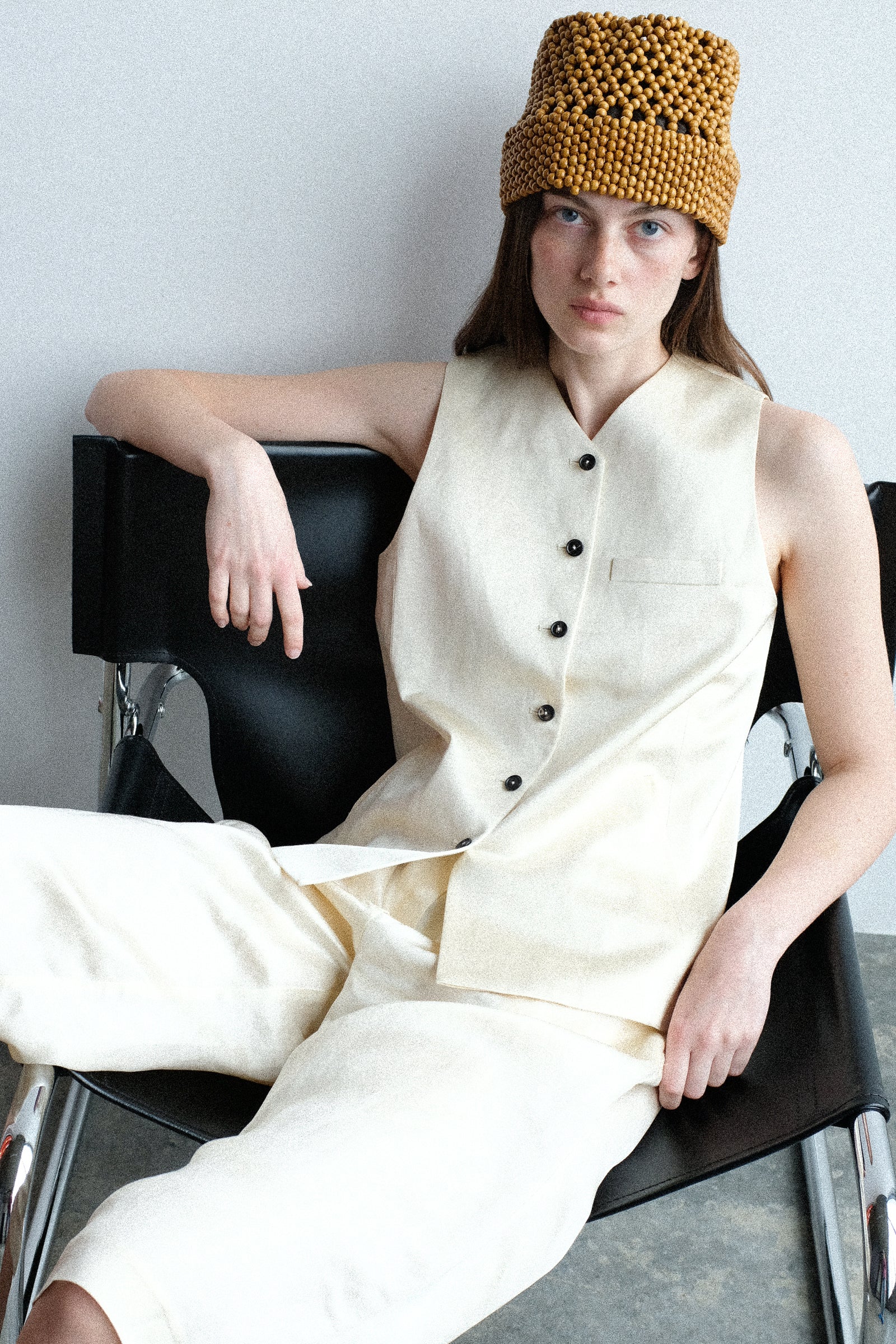 A woman sitting in a chair wearing Cordera's Wooden Beads Bucket Hat, with the Satin Tailored Vest in Marfil, and the Satin Capri Pants in Off White against a white background.