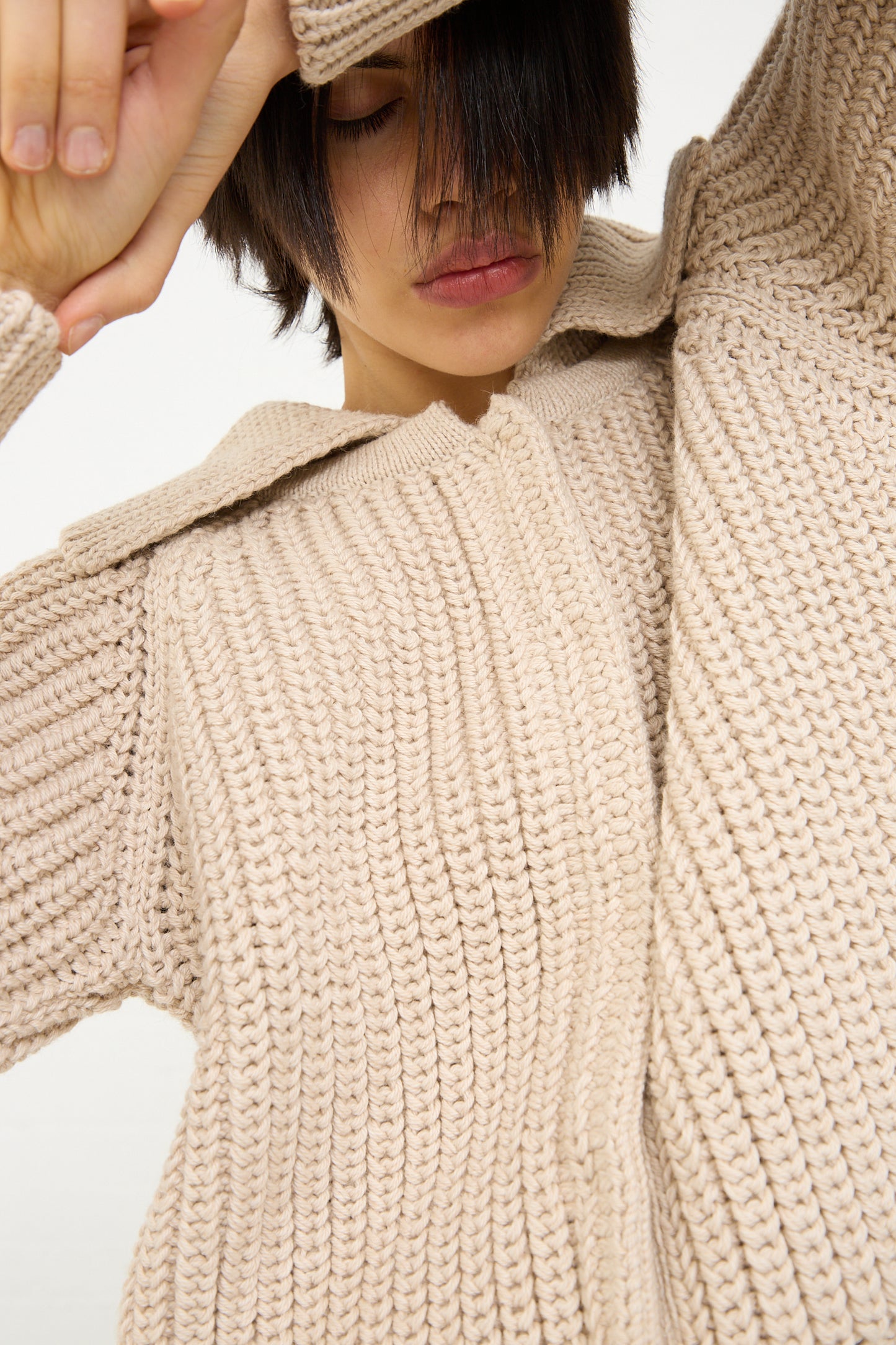 A person in a Caron Callahan Cotton and Alpaca Freda Cardigan in Ecru, with their hand resting on their forehead, partially obscuring their face.