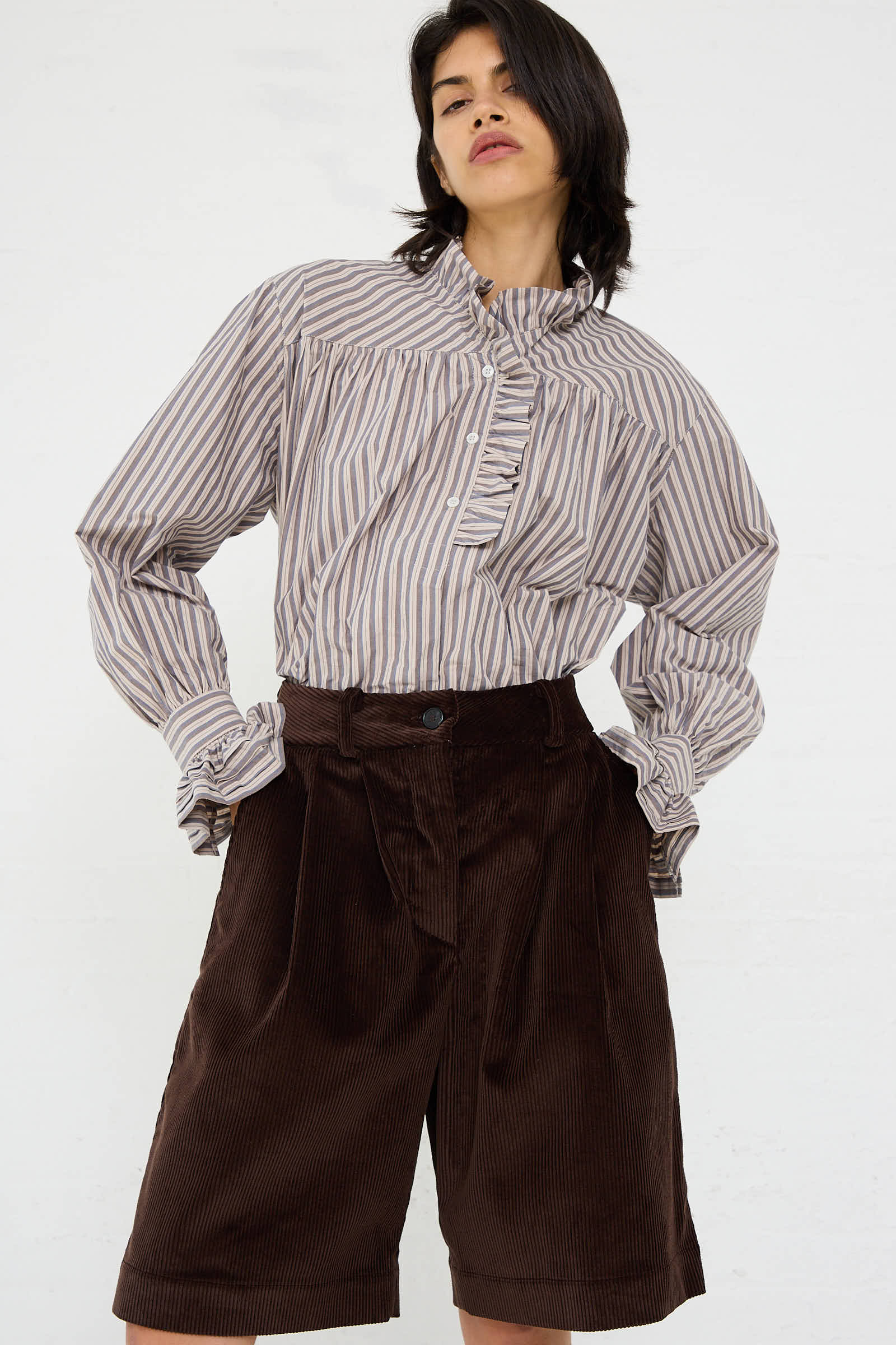 A person wearing a striped, long-sleeved blouse with a ruffled neckline and Cawley's British Cord Mara Short in Chocolate, featuring a relaxed fit. Everything is set against a plain white background.