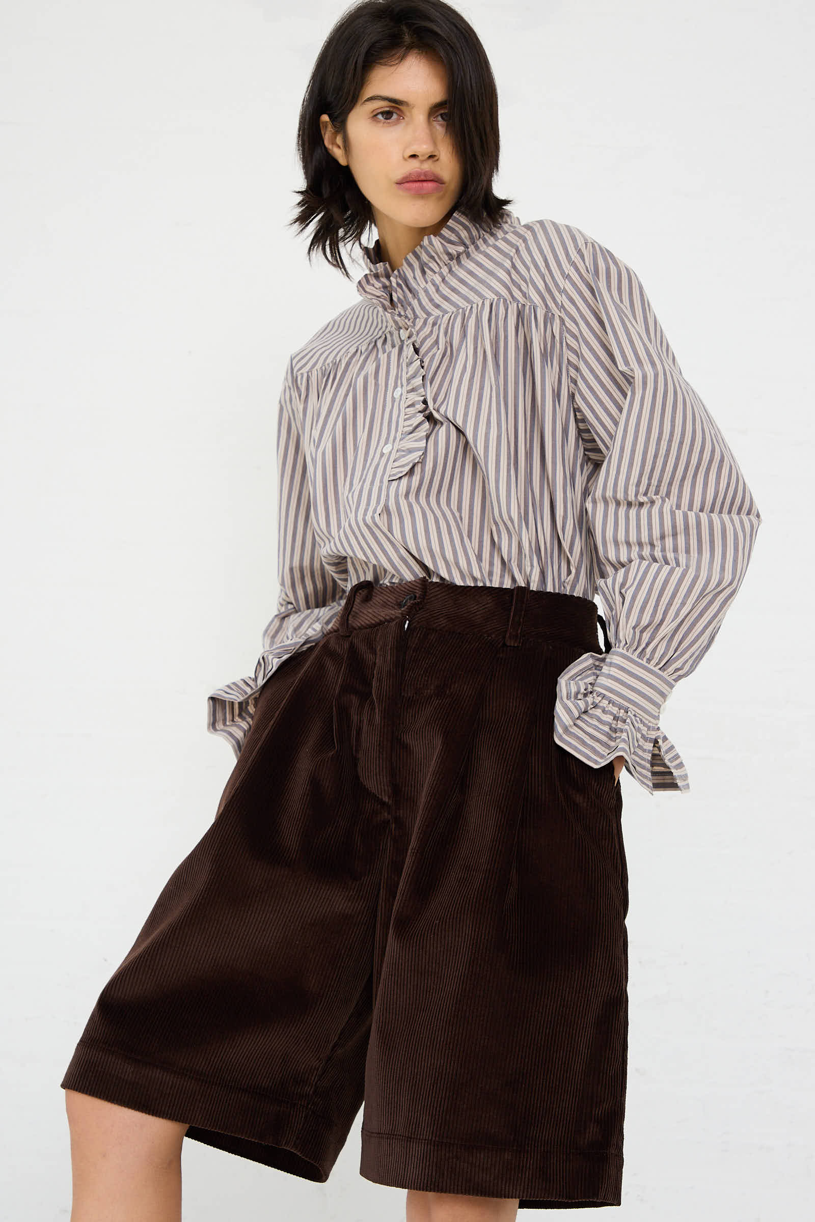 Person wearing a striped blouse with ruffled details and the British Cord Mara Short in Chocolate from Cawley, standing against a plain white background.