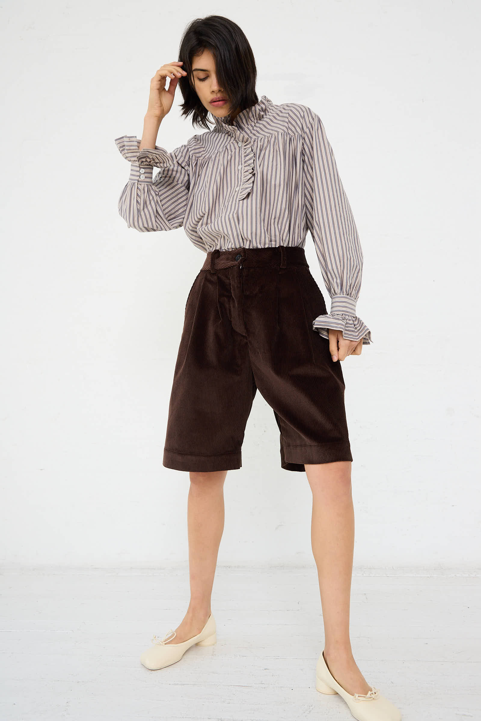 A person stands against a plain background wearing a striped blouse, Cawley's British Cord Mara Short in Chocolate, and white shoes. They have one hand near their head.