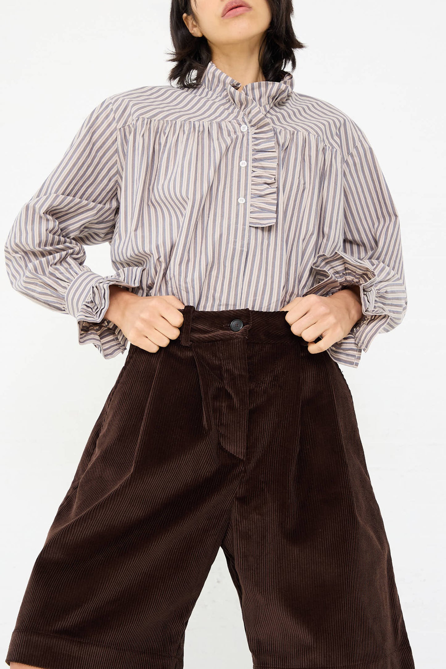 Person wearing a striped high-neck blouse with ruffled details and relaxed fit, hands on hips, paired with Cawley's British Cord Mara Short in Chocolate.