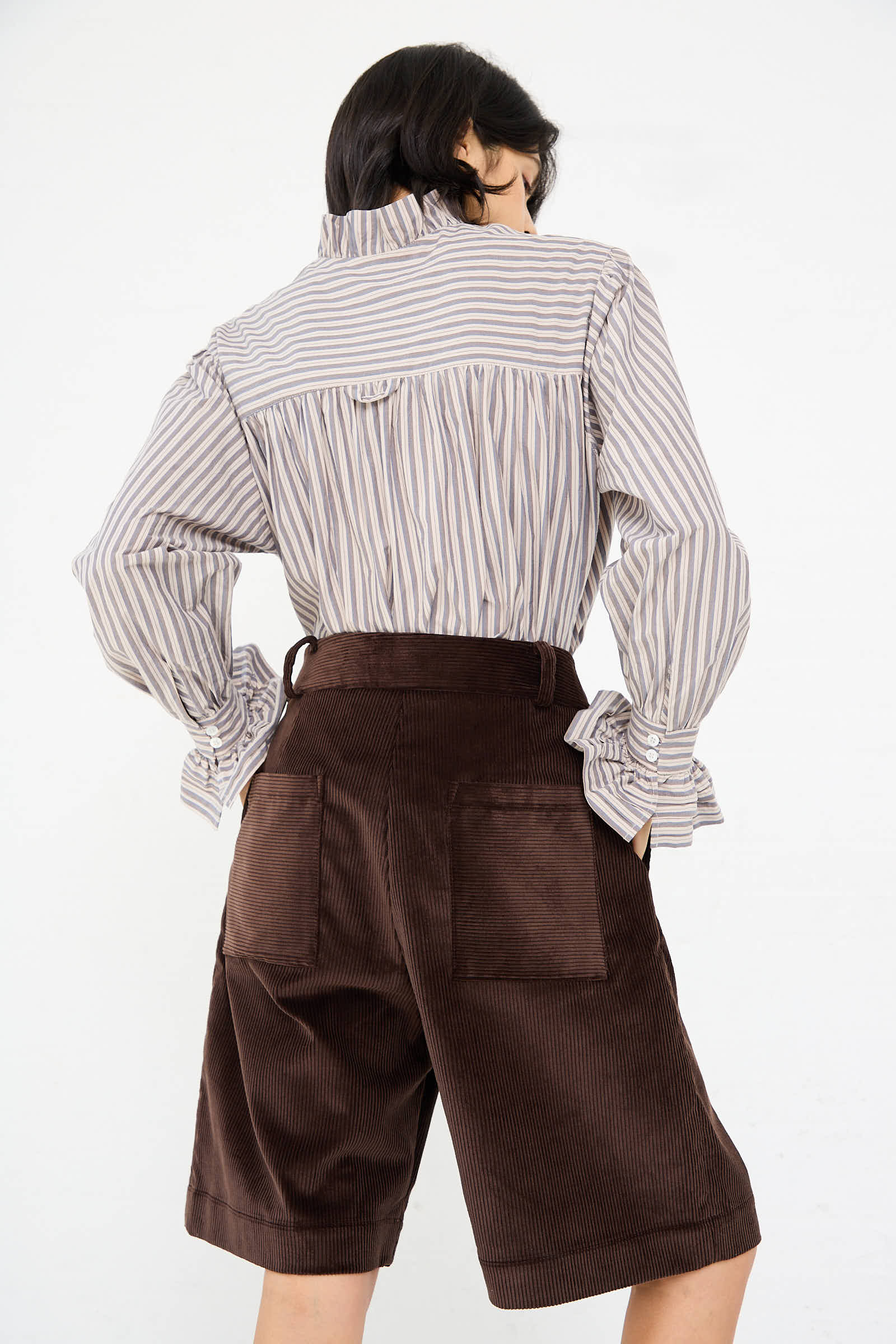 Rear view of a person wearing a striped long-sleeve shirt and high-waisted Cawley British Cord Mara Shorts in Chocolate, made from 100% cotton, standing against a plain white background.