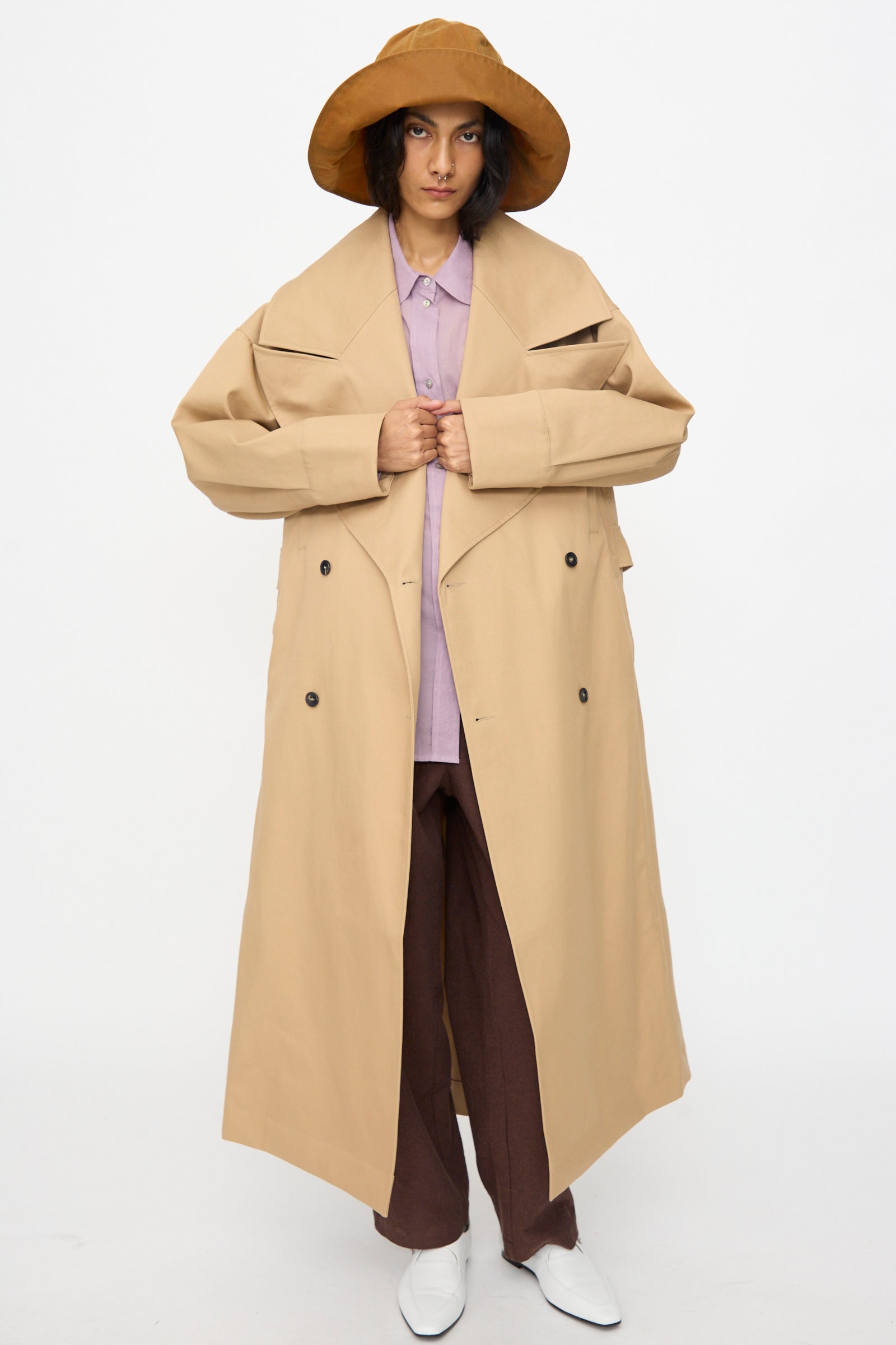 A person wears a wide-brimmed brown hat, the Cawley British Cotton Gabardine Agnes Mac in Nutmeg, a lavender shirt, brown pants, and white shoes while standing against a plain white background.
