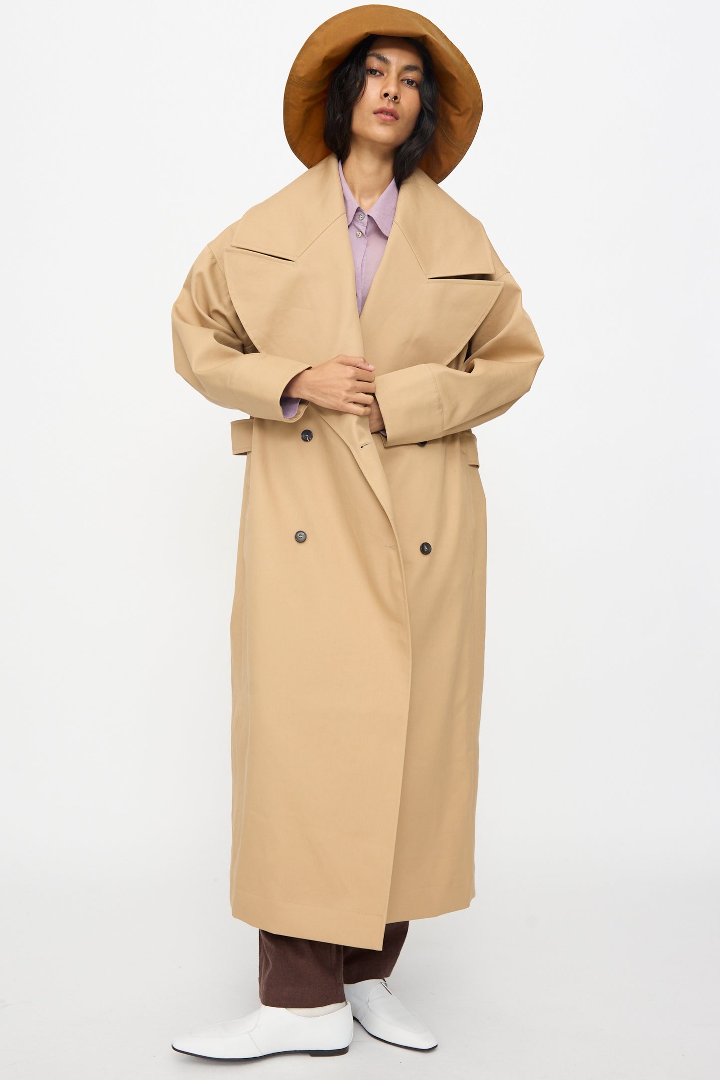 A person stands against a plain white background wearing a wide-brimmed tan hat, light purple shirt, brown pants, white shoes, and the British Cotton Gabardine Agnes Mac in Nutmeg by Cawley.