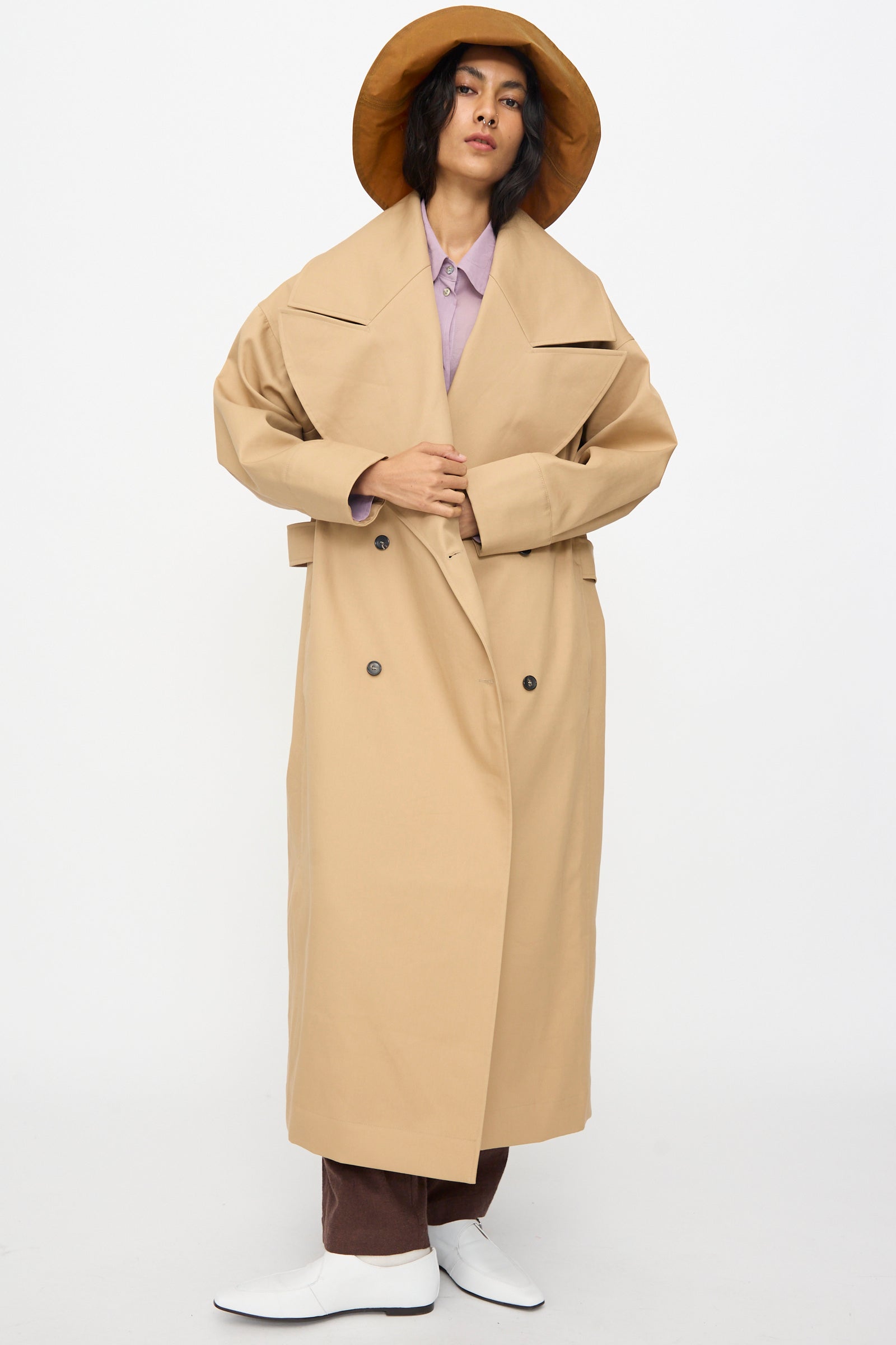 A person stands against a plain white background wearing a wide-brimmed tan hat, light purple shirt, brown pants, white shoes, and the British Cotton Gabardine Agnes Mac in Nutmeg by Cawley.